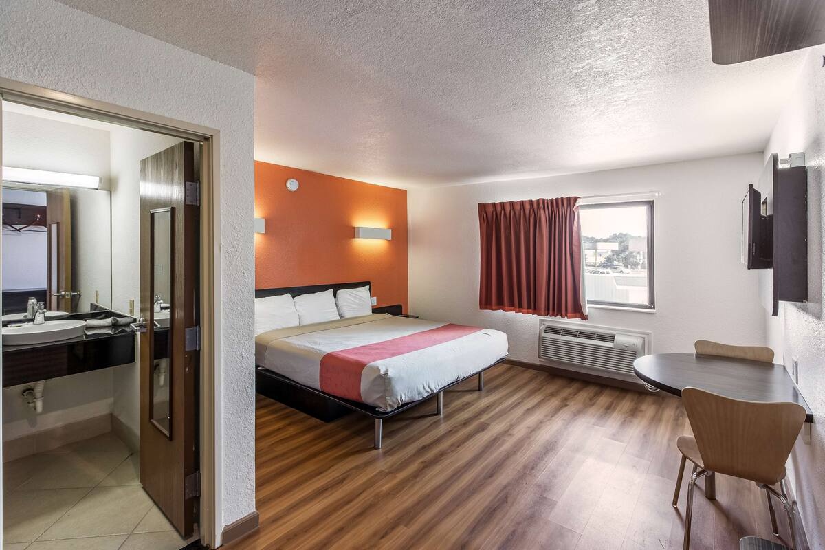 A hotel room with a bed, a desk, a chair, a window with curtains, a mirror, and a bathroom with a sink and mirror.