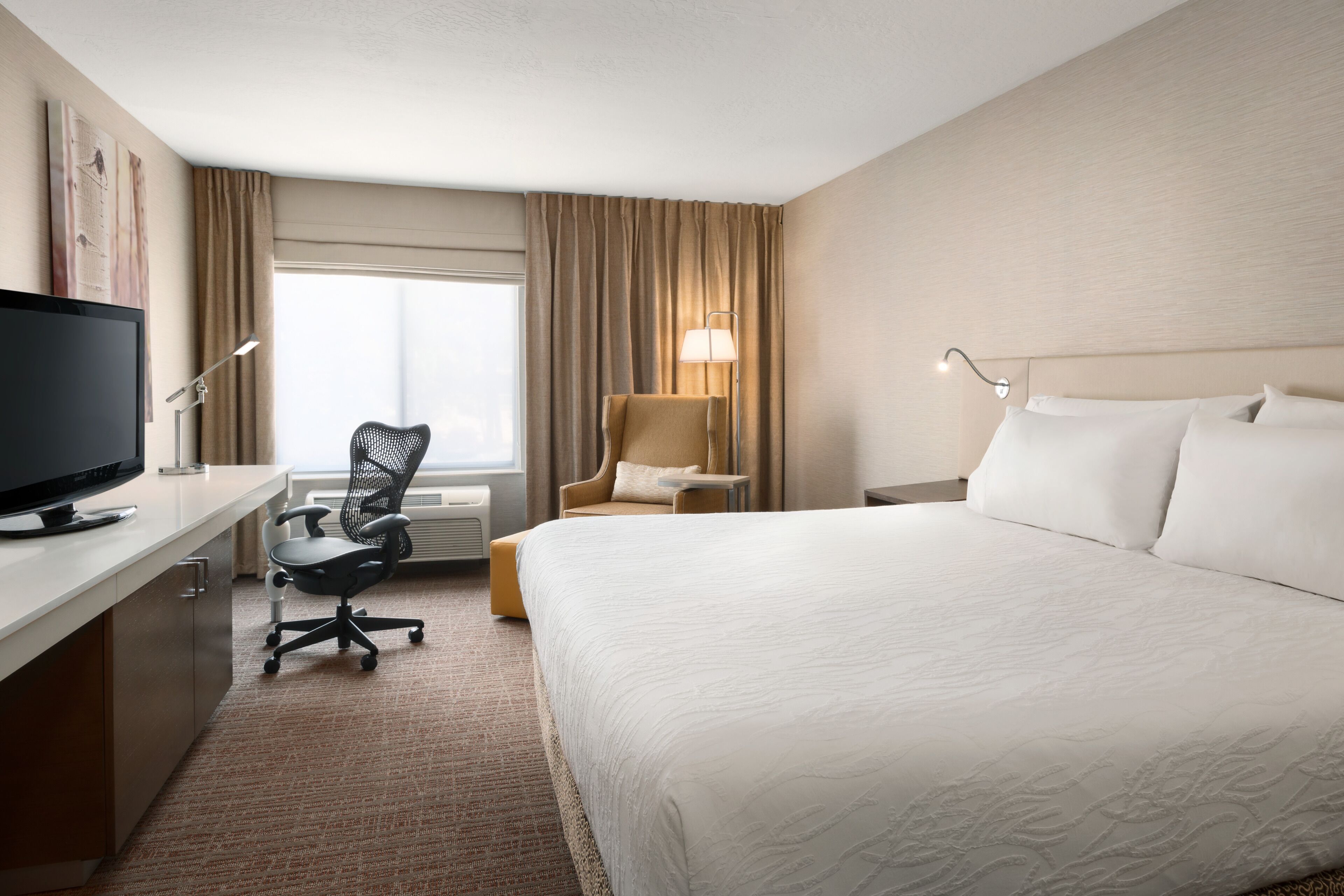A hotel room with a large bed, a desk with a flat-screen TV, a chair, a lamp, and a window with curtains.