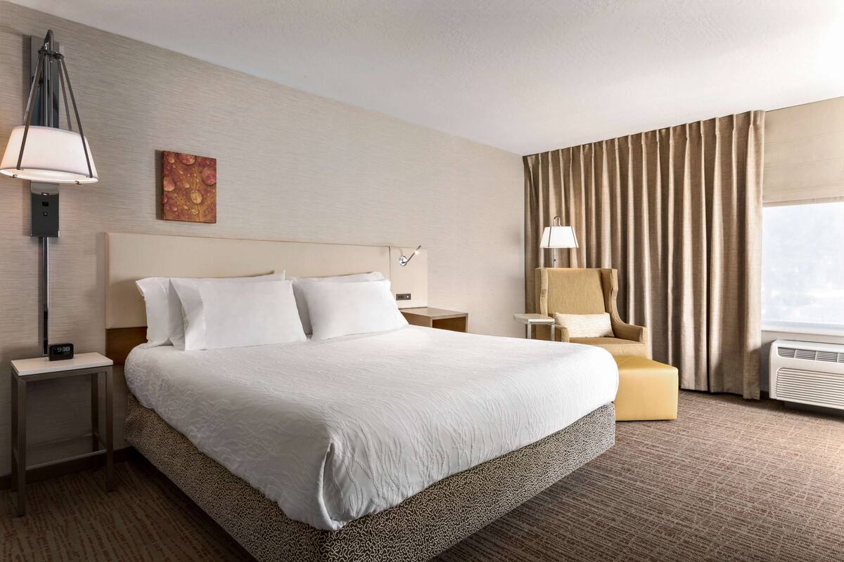 A hotel room with a large bed, a bedside table with a lamp, a chair, and a window with curtains.