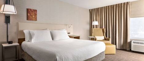 A hotel room with a large bed, a bedside table with a lamp, a chair, and a window with curtains.