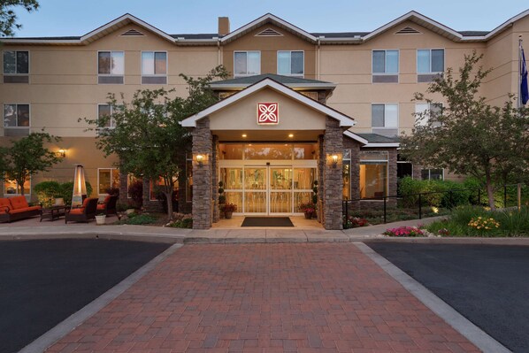 A hotel entrance with a brick pathway leading to a glass door, stone pillars, and a landscaped garden with potted plants and a seating area.