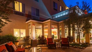 An outdoor patio area with seating and a glass table, surrounded by plants and a building with large windows. - Hilton Garden Inn - Flagstaff (Flagstaff)