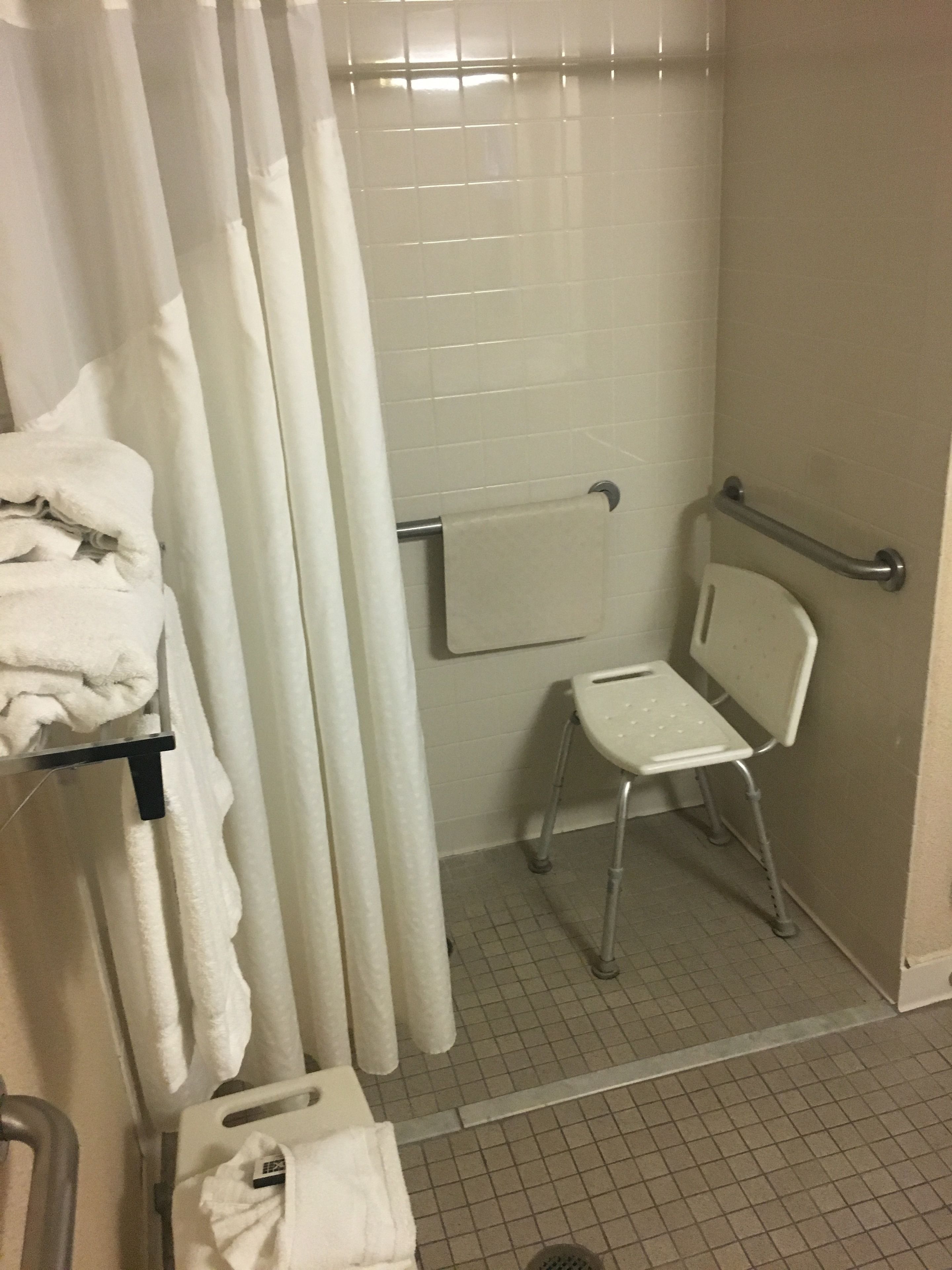 A bathroom with a shower, a toilet, a grab bar, and a chair.