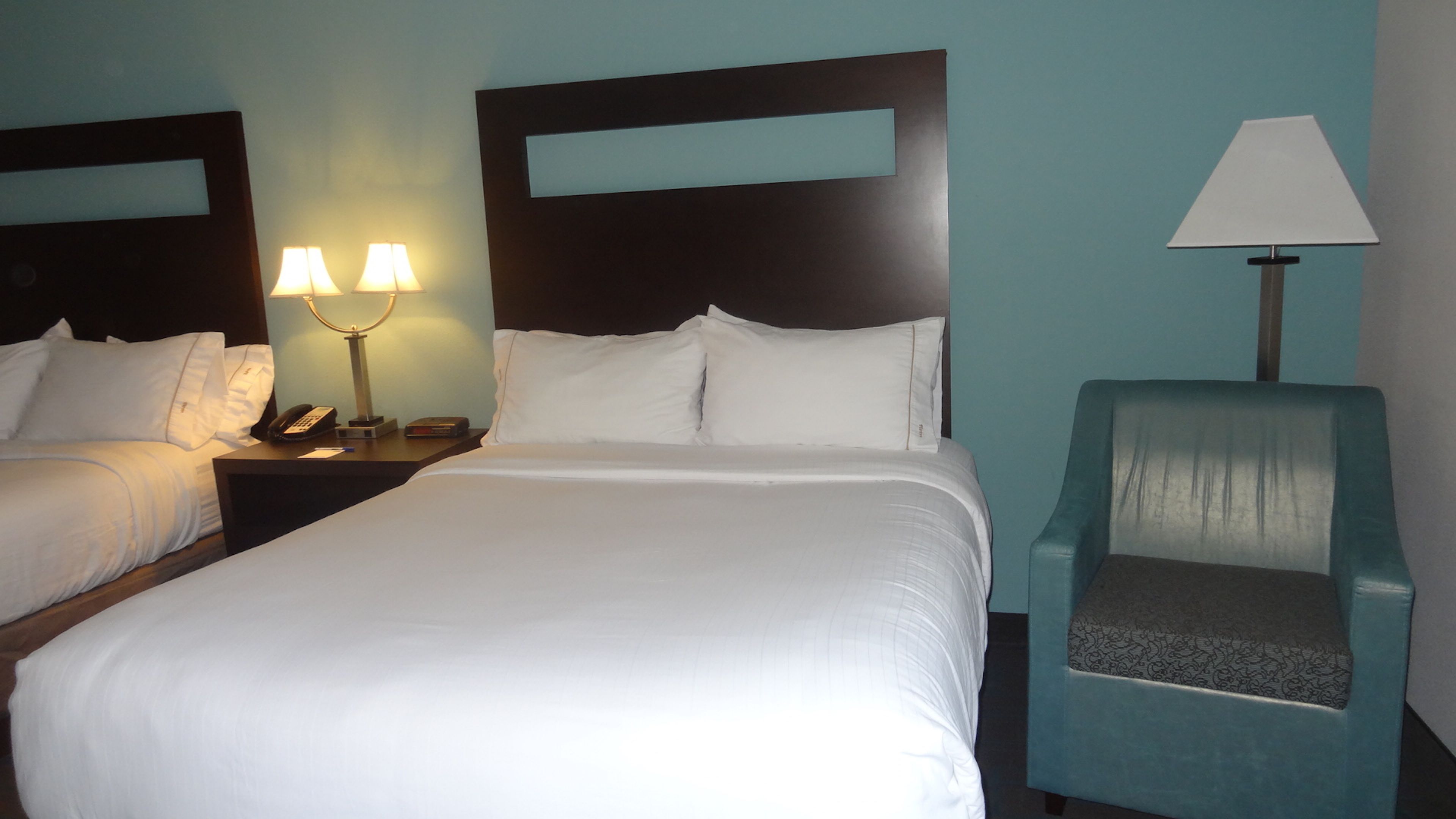 A hotel room with two beds, a nightstand with a lamp, a chair, and a wall-mounted TV.