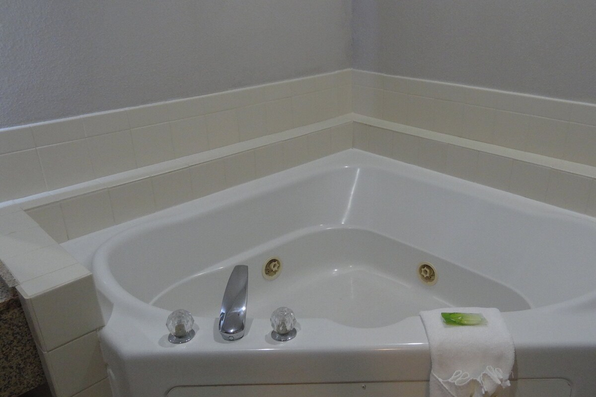 Una bañera en esquina con un grifo y dos mandos, una toalla al lado y un objeto verde en el borde.