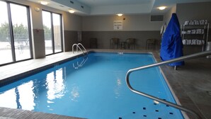 Une piscine intérieure avec un panneau indiquant qu’il est interdit de plonger, une terrasse en bois et un espace couvert où se trouvent des serviettes.