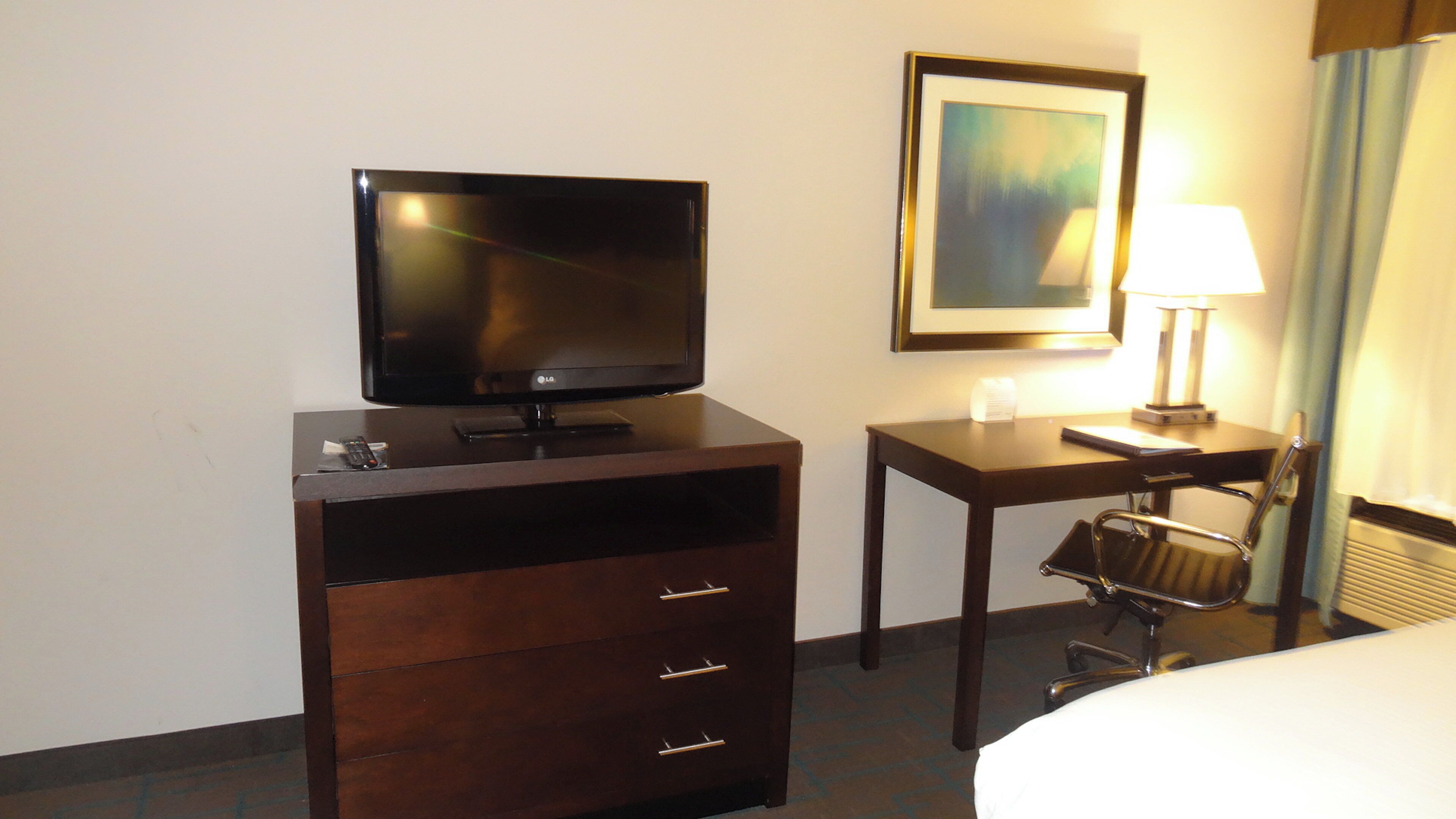 Une chambre d’hôtel avec un téléviseur à écran plat posé sur une commode en bois, un bureau avec une lampe, une chaise et un lit.