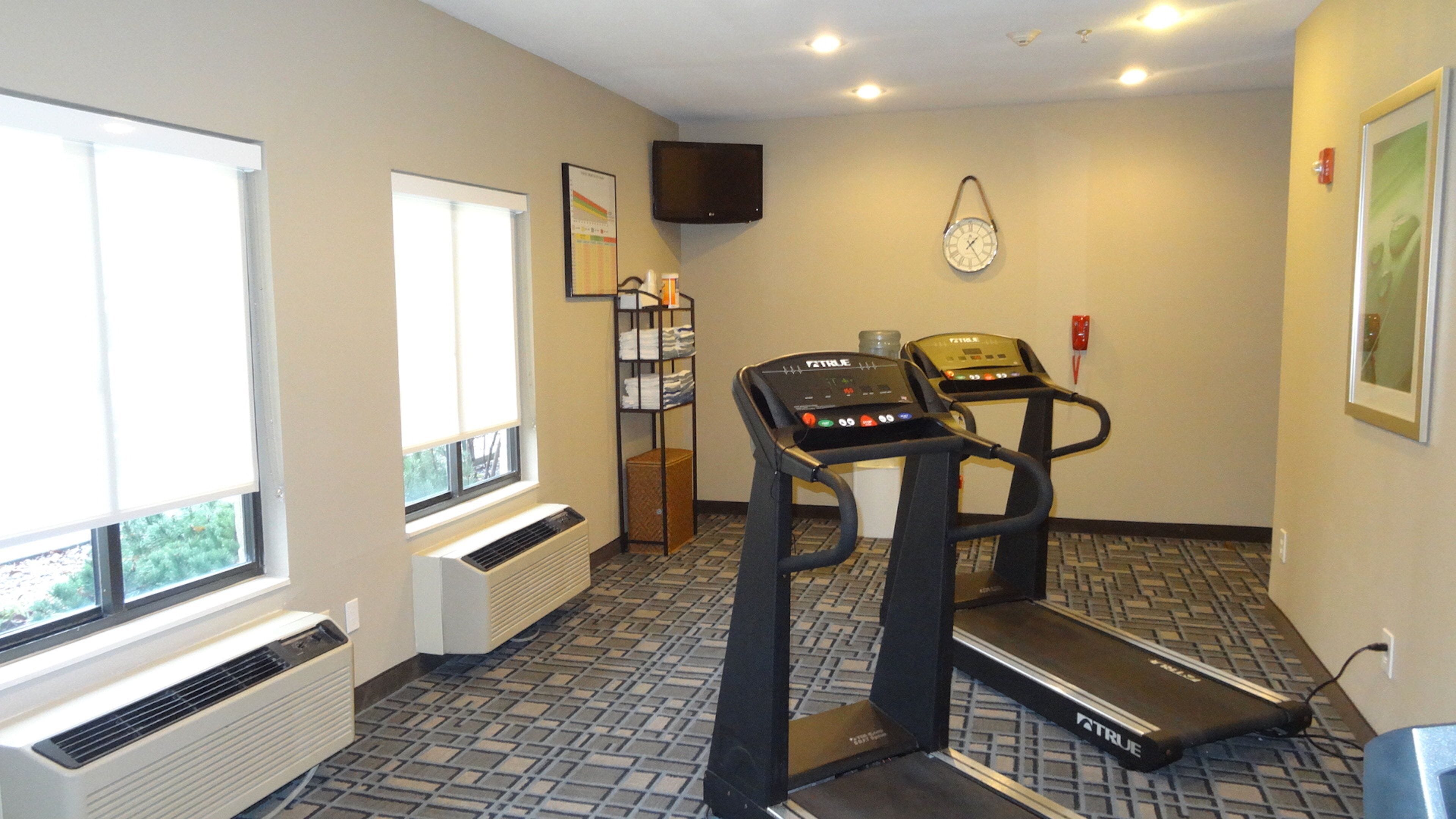 Un gimnasio con una cinta de correr, un televisor de pared y una ventana con persianas.