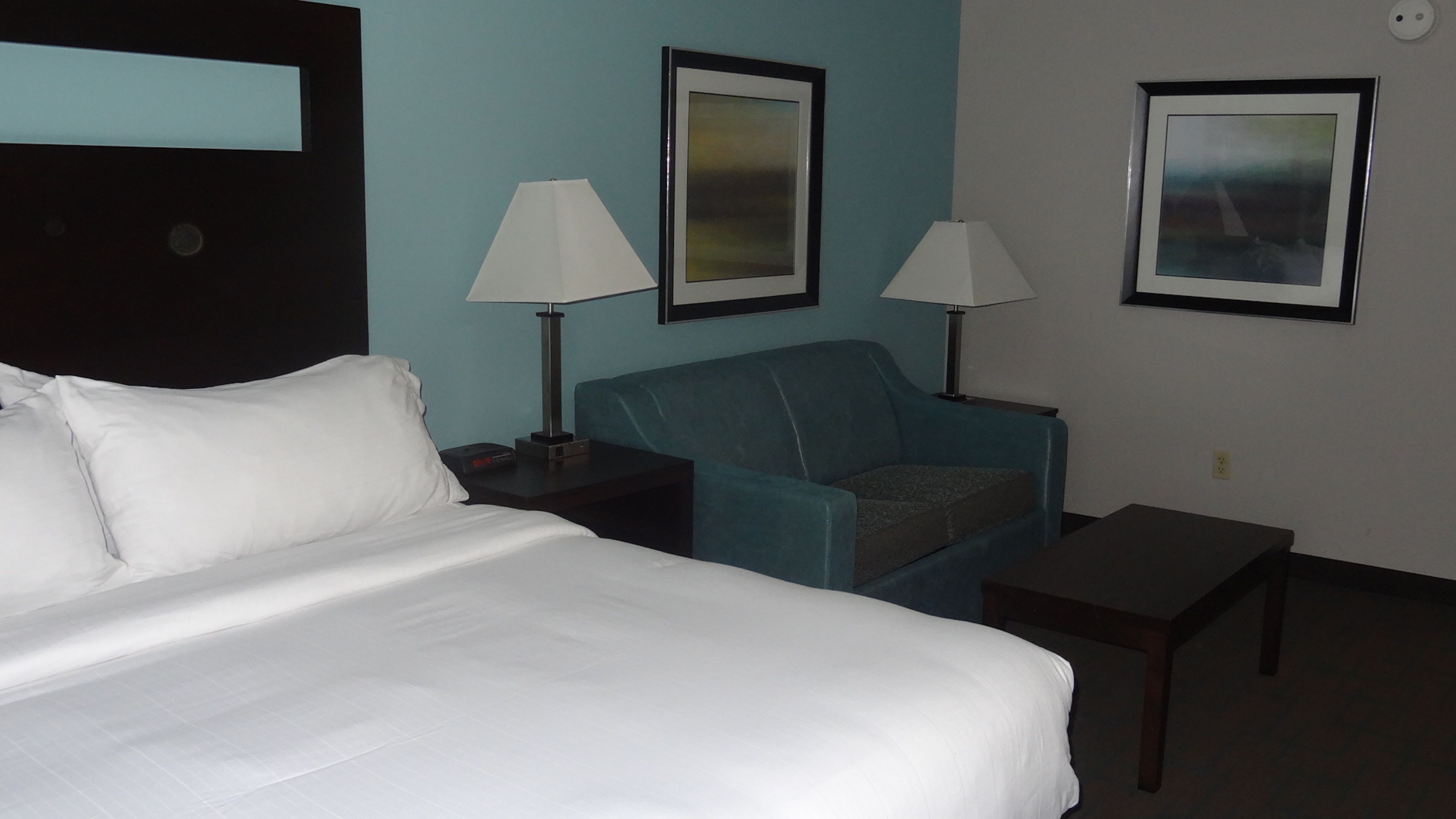 A hotel room with a bed, a blue armchair, two bedside tables with lamps, and two framed pictures on the wall.