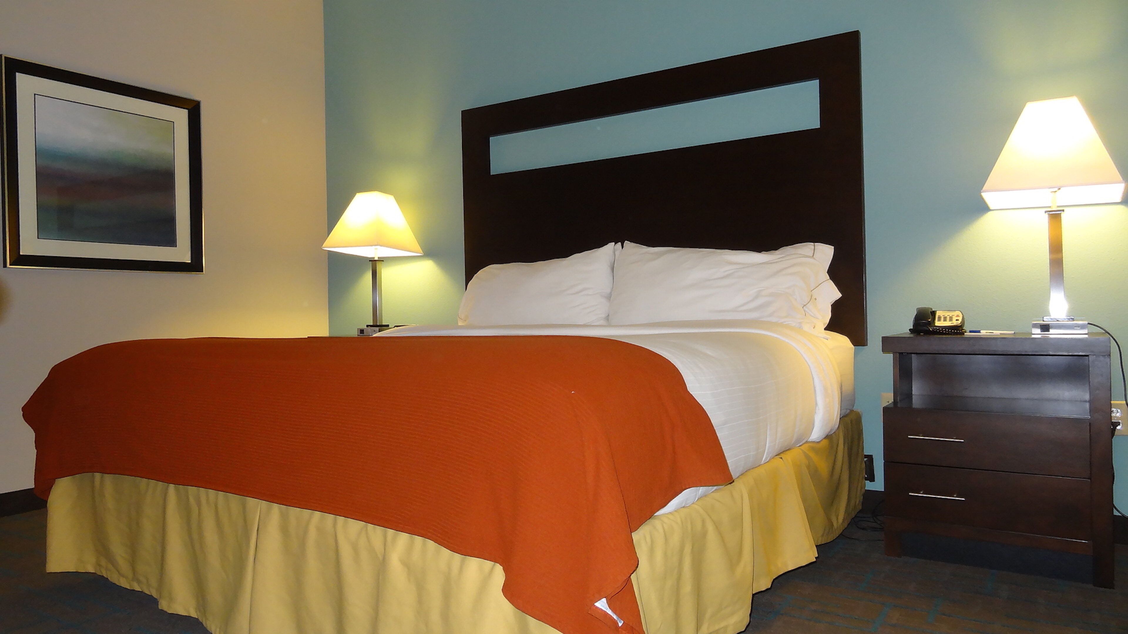 Habitación de hotel con cama, dos lámparas de noche, una mesita de noche y un cuadro enmarcado en la pared.