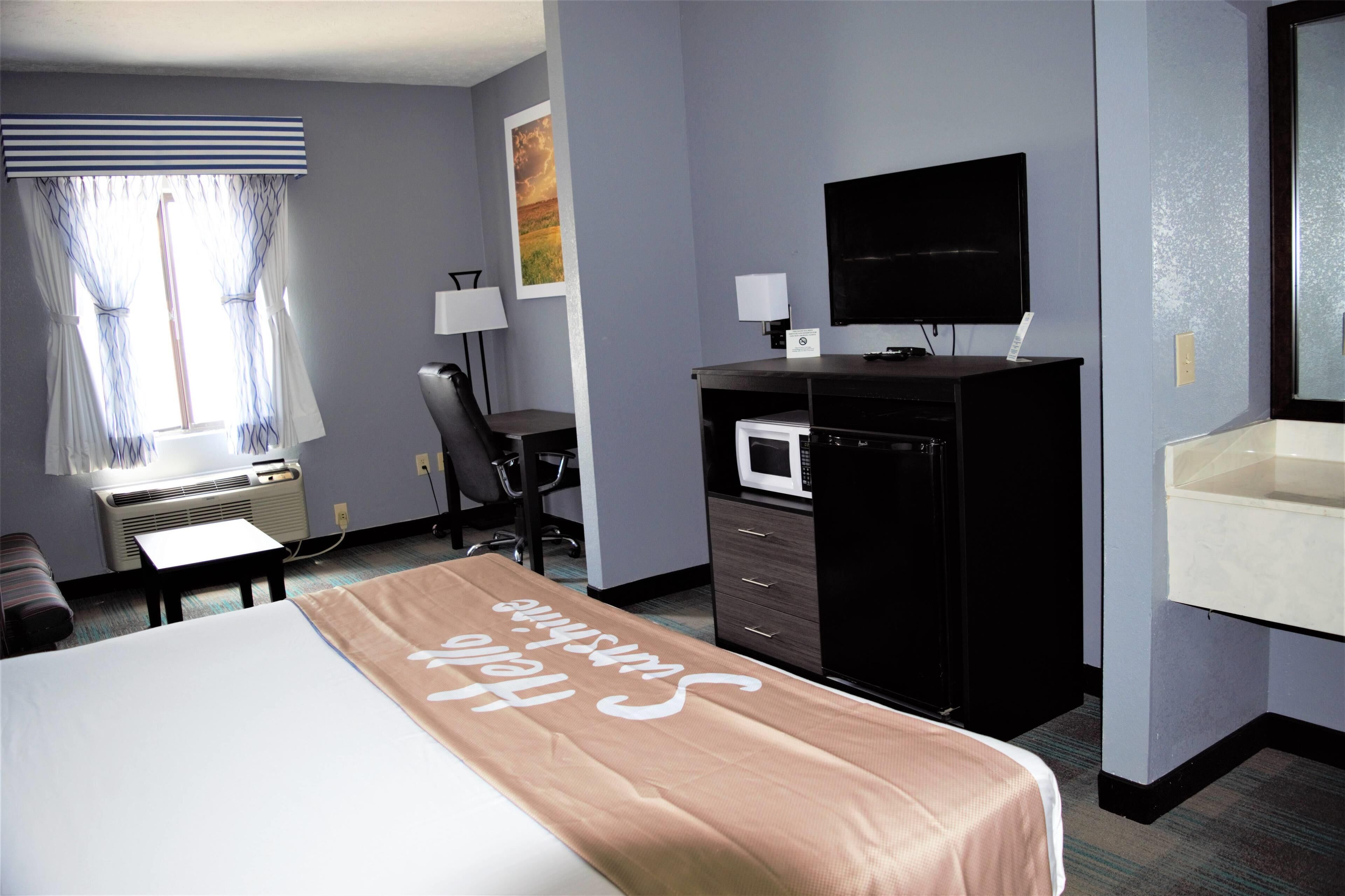 A hotel room with a bed, a desk with a computer, a microwave, a TV, and a bathroom with a sink and mirror.