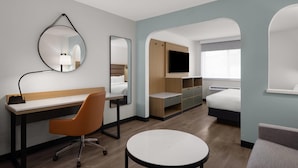Habitación de hotel con escritorio, silla, mesa redonda, cama, televisor y espacio de almacenamiento integrado.