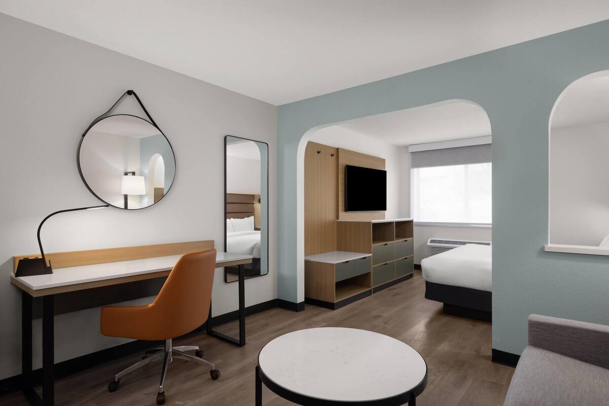 A hotel room with a desk, chair, round table, bed, TV, and built-in storage.