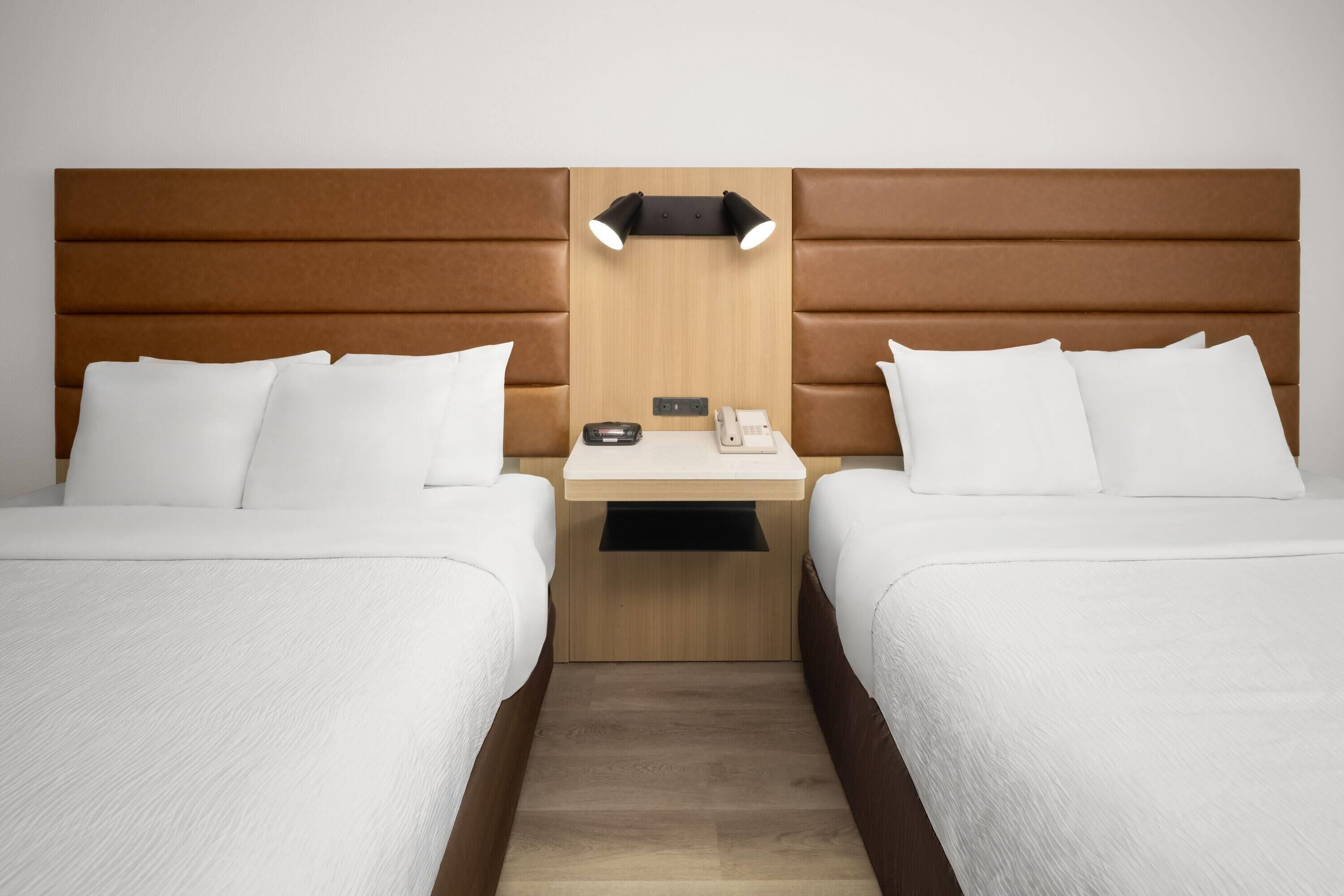 Quarto de hotel com duas camas, cabeceira de madeira, mesa de cabeceira com abajur e telefone.