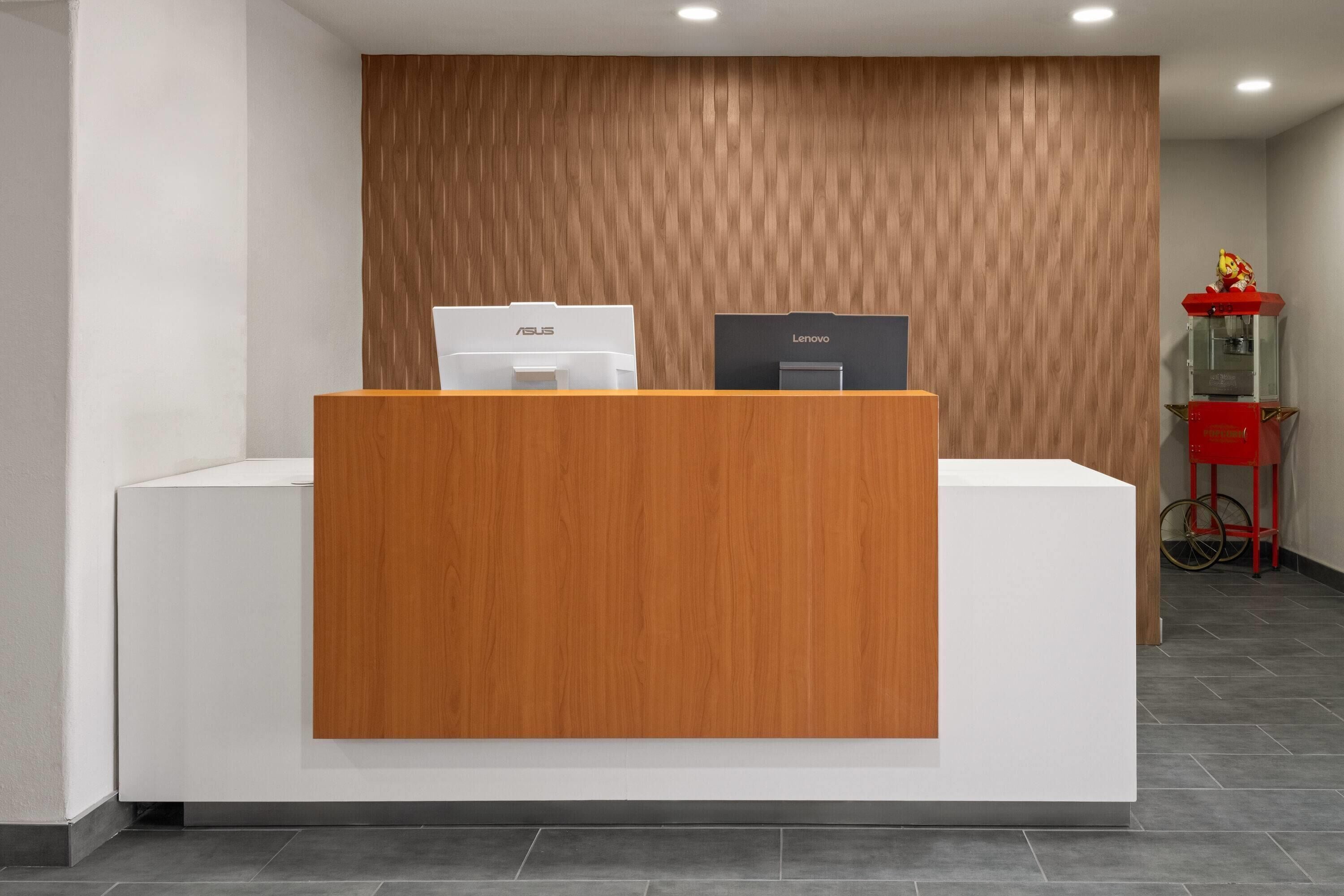Un mostrador de recepción con un panel frontal de madera, una computadora ASUS y una Lenovo encima, y una máquina expendedora roja al fondo.