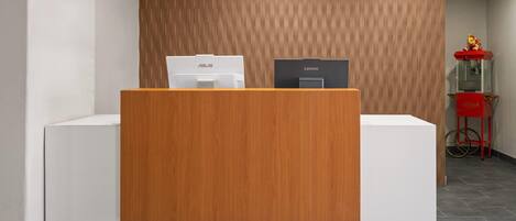 Un mostrador de recepción con un panel frontal de madera, una computadora ASUS y una Lenovo encima, y una máquina expendedora roja al fondo.
