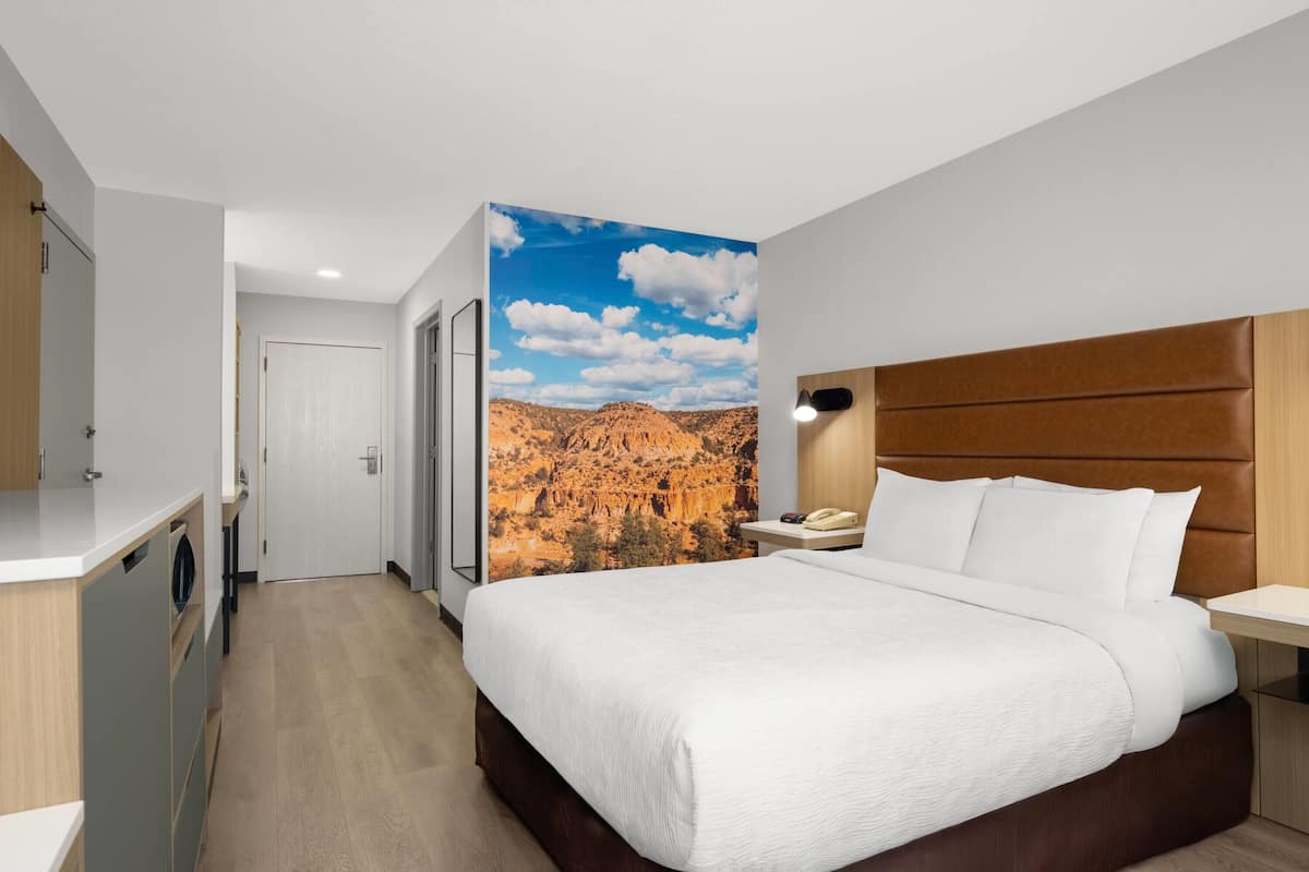 A hotel room with a bed, a nightstand, a wall-mounted lamp, a wardrobe, and a door leading to a bathroom.