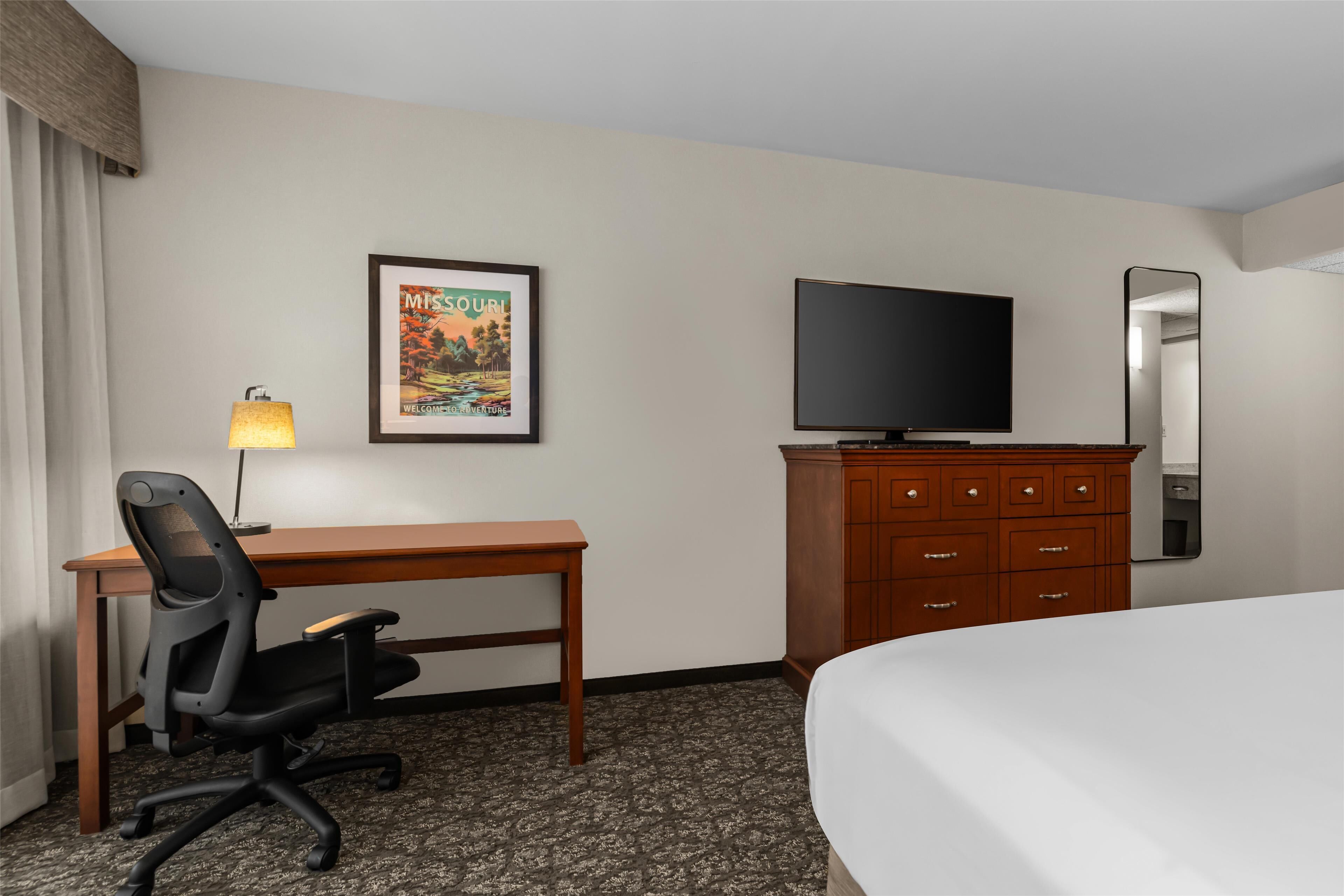 Una habitación de hotel con cama, escritorio con silla, televisor, cómoda, espejo y un cuadro en la pared.