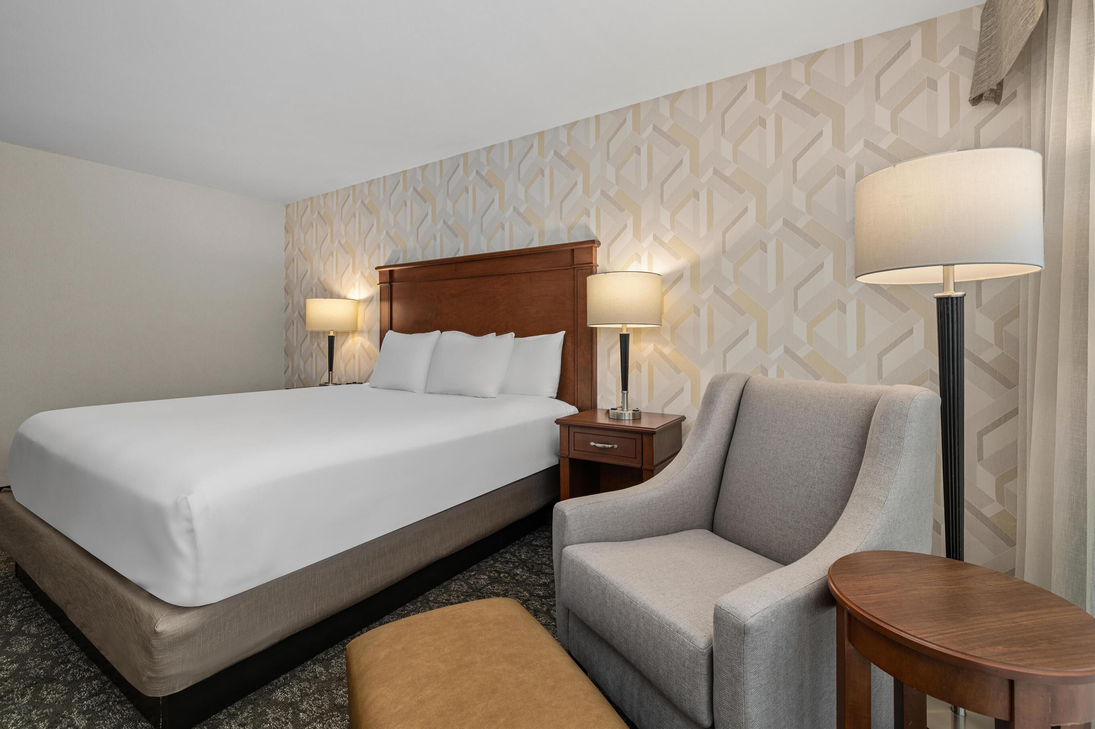 Habitación de hotel con una cama grande, dos lámparas de noche, un sillón gris, una pequeña mesa de madera y un papel tapiz estampado.
