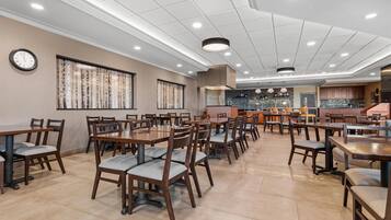 A spacious dining area with multiple tables and chairs, a kitchen in the background, and a clock on the wall.