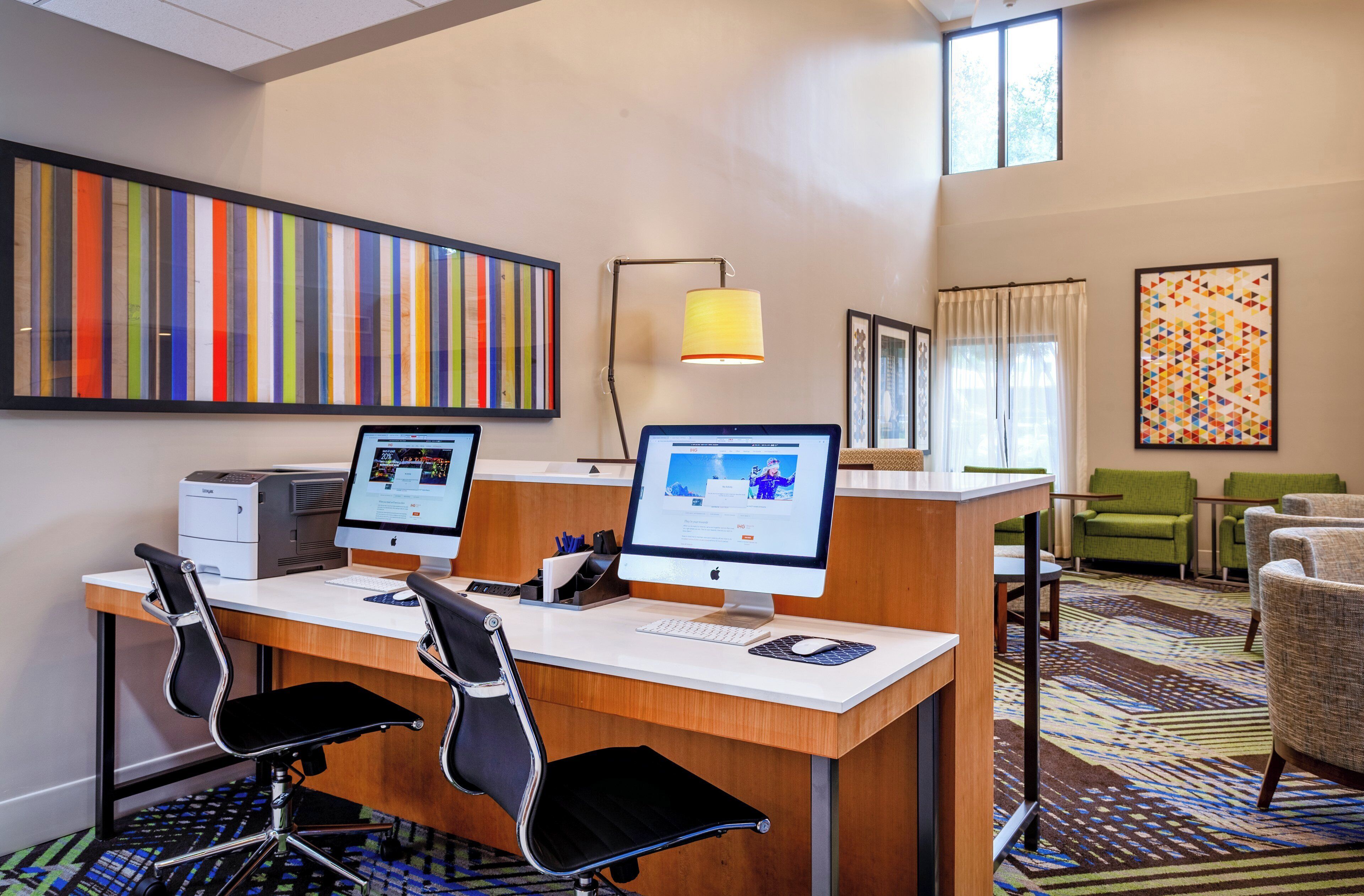 A hotel room with two desktop computers, a printer, a desk, two chairs, a lamp, and a large window.
