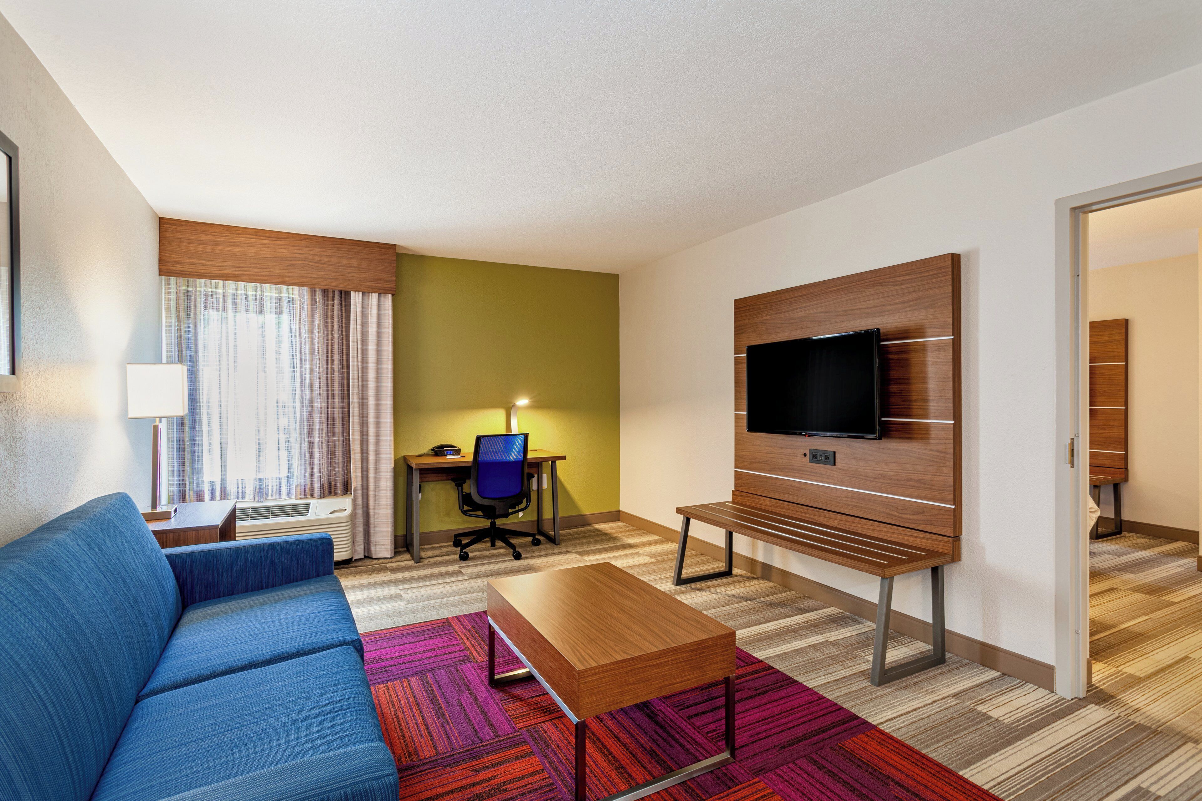 Une chambre d’hôtel avec un canapé bleu, une table basse en bois, un bureau avec une chaise, un téléviseur à écran plat fixé sur un panneau en bois et une moquette à motifs.