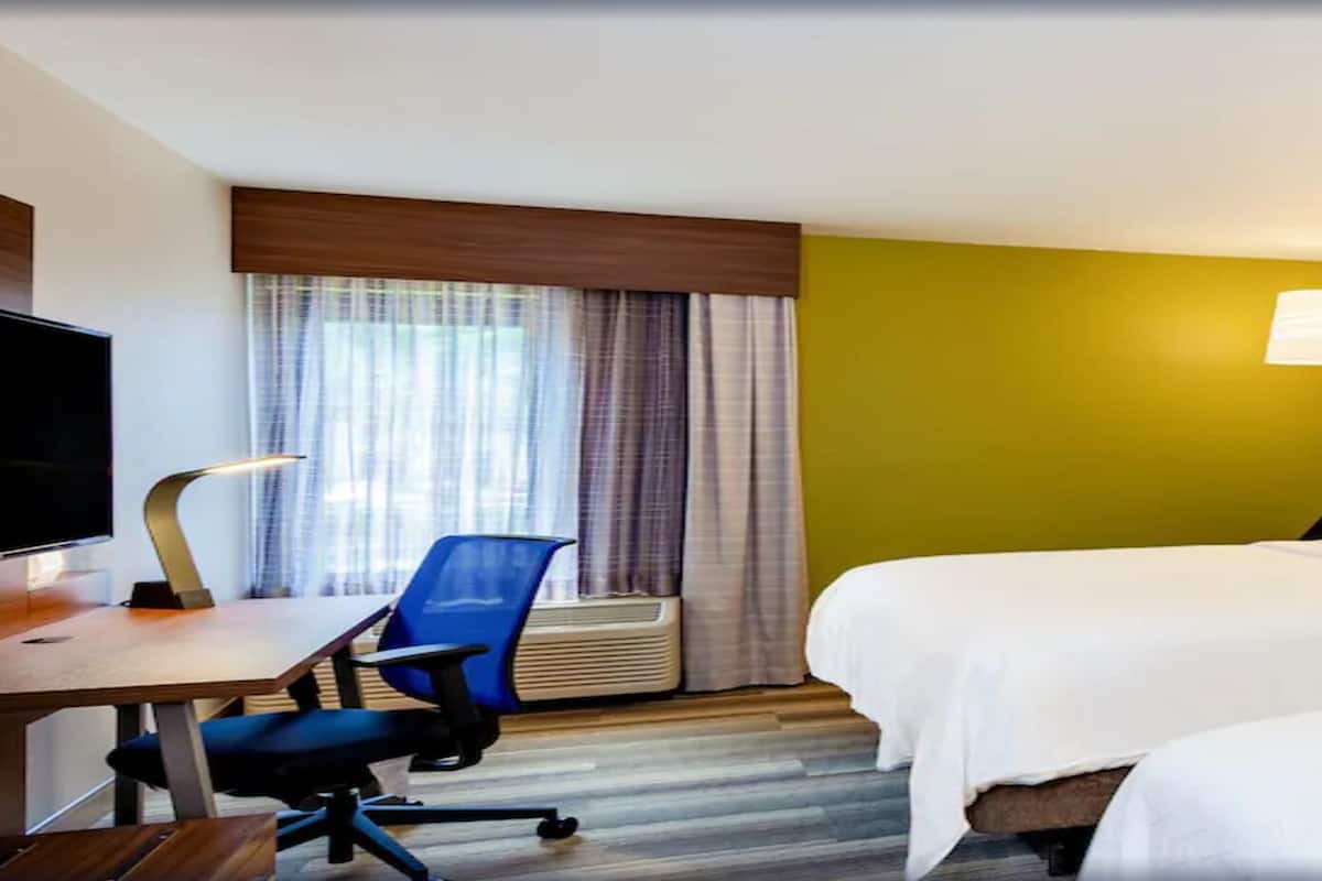 Ein Hotelzimmer mit zwei Betten, einem Schreibtisch mit Stuhl, einem Fernseher und einem Fenster mit Vorhängen.
