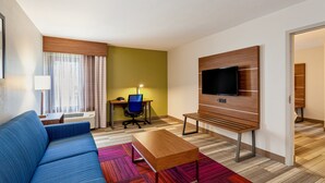 Une chambre d’hôtel moderne avec un canapé bleu, une table basse en bois, un téléviseur fixé au mur, un bureau avec une chaise et une moquette colorée.