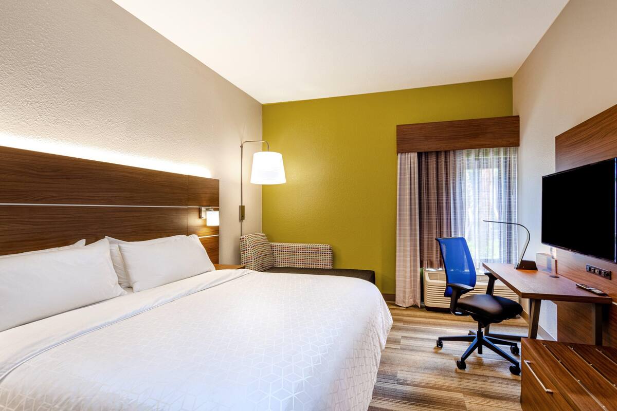 Ein Hotelzimmer mit einem großen Bett, einem Schreibtisch mit Stuhl, einem Fernseher und einem Fenster mit Vorhängen.