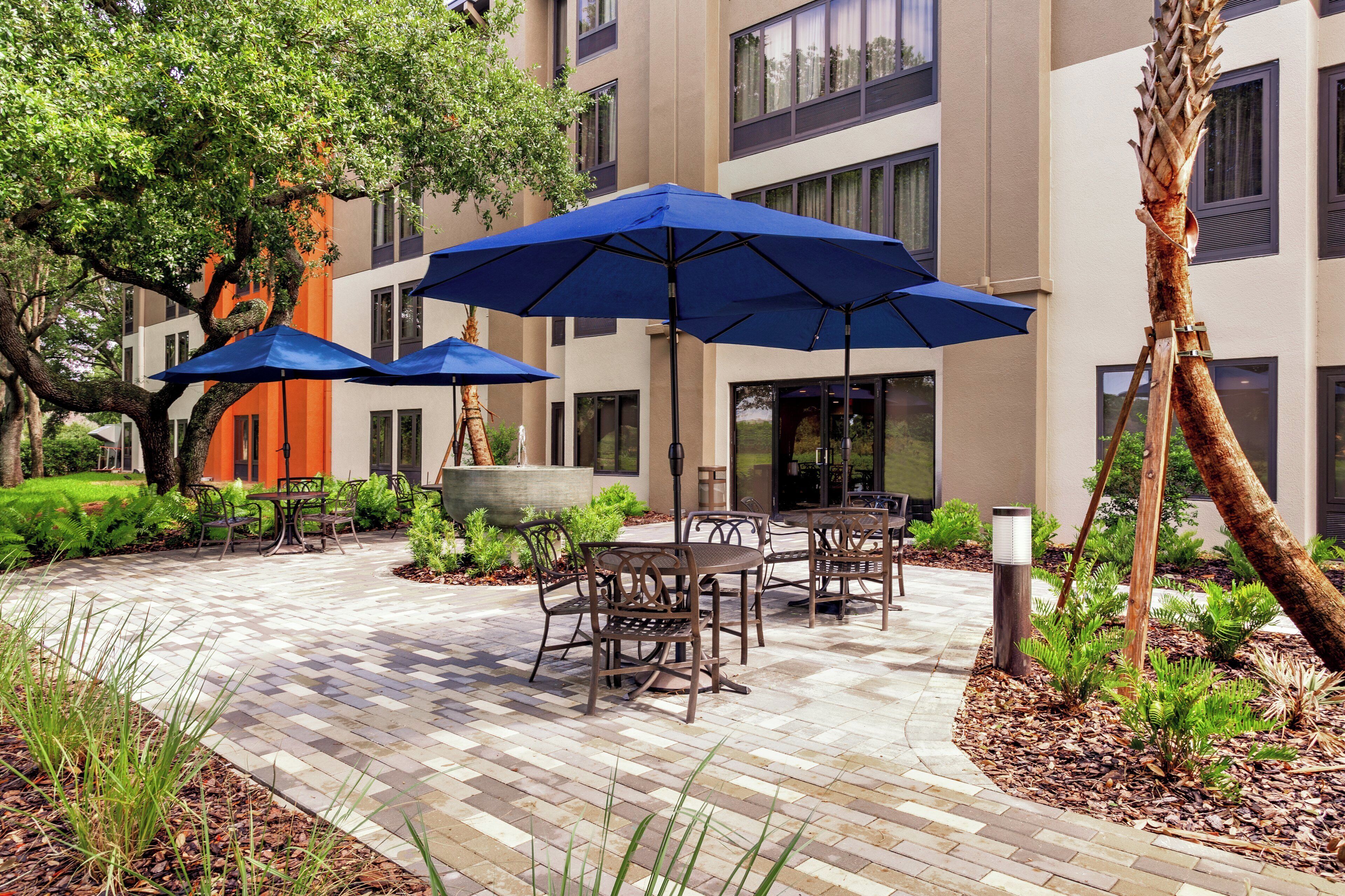 An outdoor patio area with tables and chairs, blue umbrellas, and a building with multiple windows.
