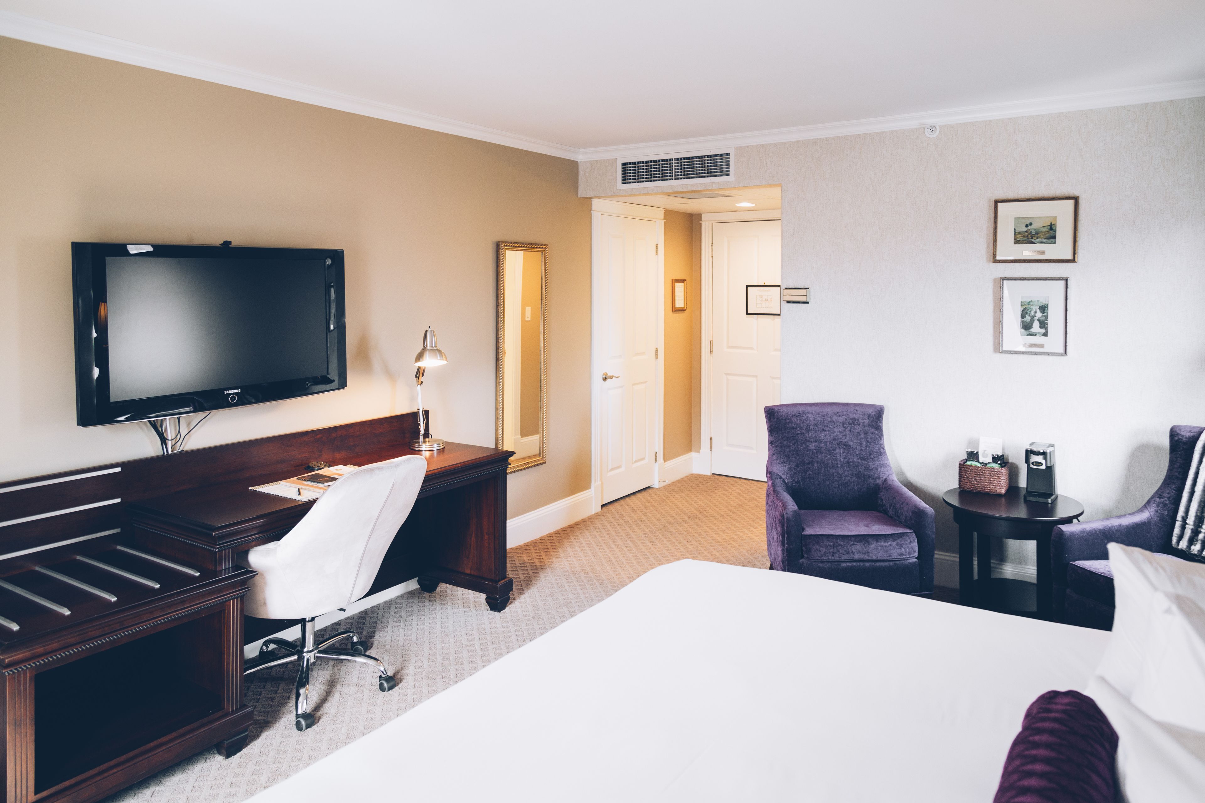 Quarto de hotel com cama, escrivaninha com cadeira, televisão e mesinha de cabeceira com abajur.