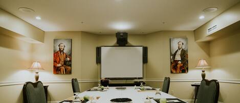 A conference room with a long table, chairs, a projector screen, and two paintings on the walls.