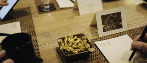 A wooden table with a black mug, a bowl of gold-colored items, a business card, and a pen.