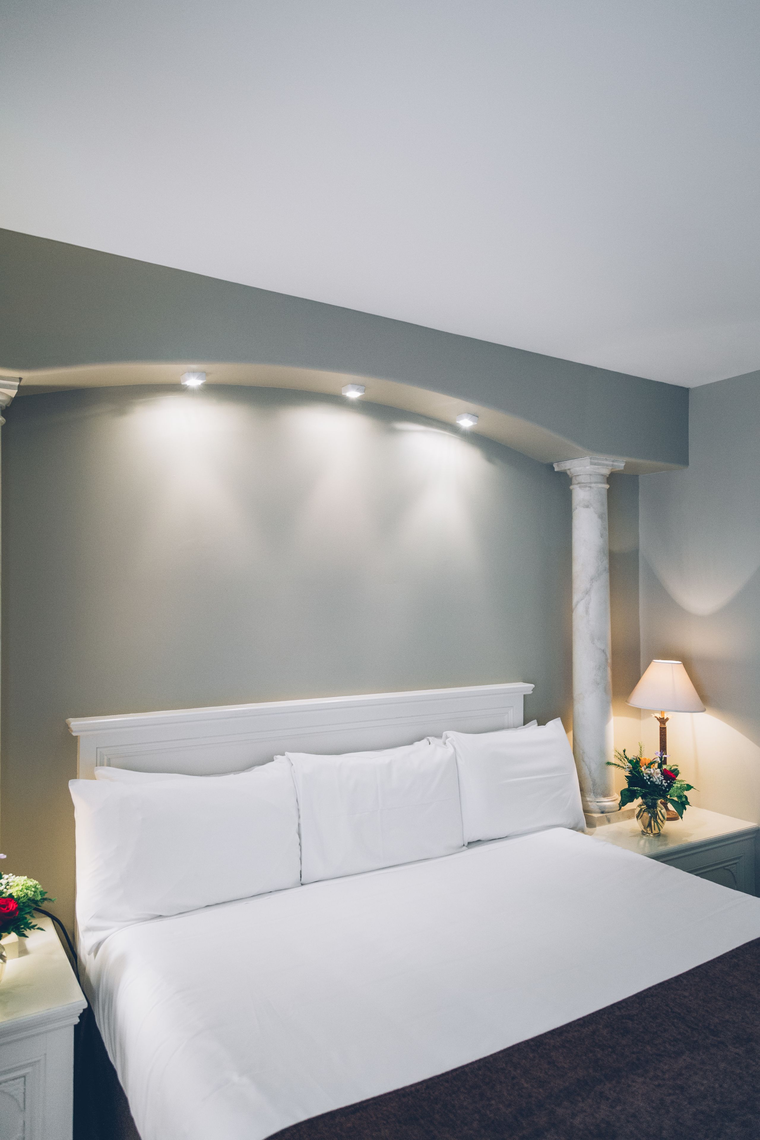 Una cama bien hecha con sábanas blancas, una mesita de noche con una lámpara y un florero, y una columna al fondo.