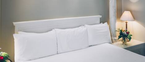 A neatly made bed with white linens, a nightstand with a lamp and a vase of flowers, and a column in the background.