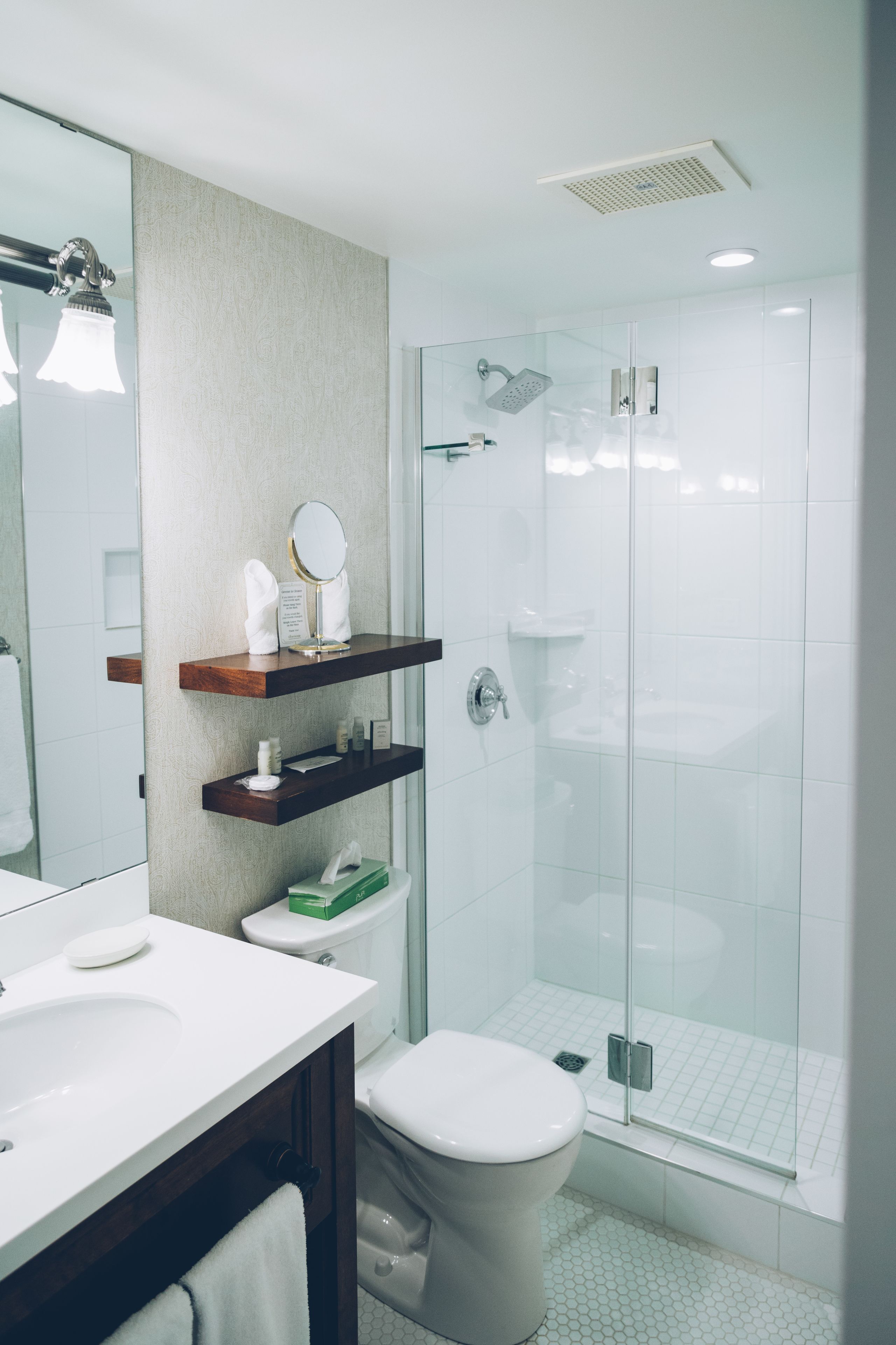 A modern bathroom with a glass shower enclosure, a white toilet, a vanity with a mirror, and a shelf with toiletries.