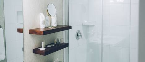 A modern bathroom with a glass shower enclosure, a white toilet, a vanity with a mirror, and a shelf with toiletries.