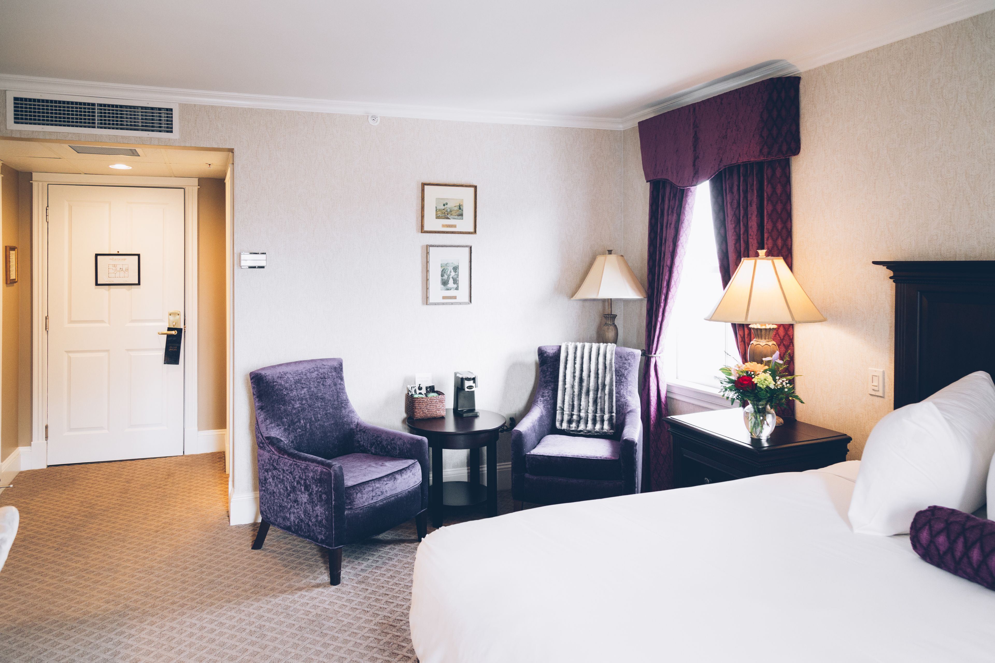 Una habitación de hotel con una cama, dos sillones, una mesita de noche con una lámpara, una pequeña mesa con una cesta y un marco de fotos, y una puerta.