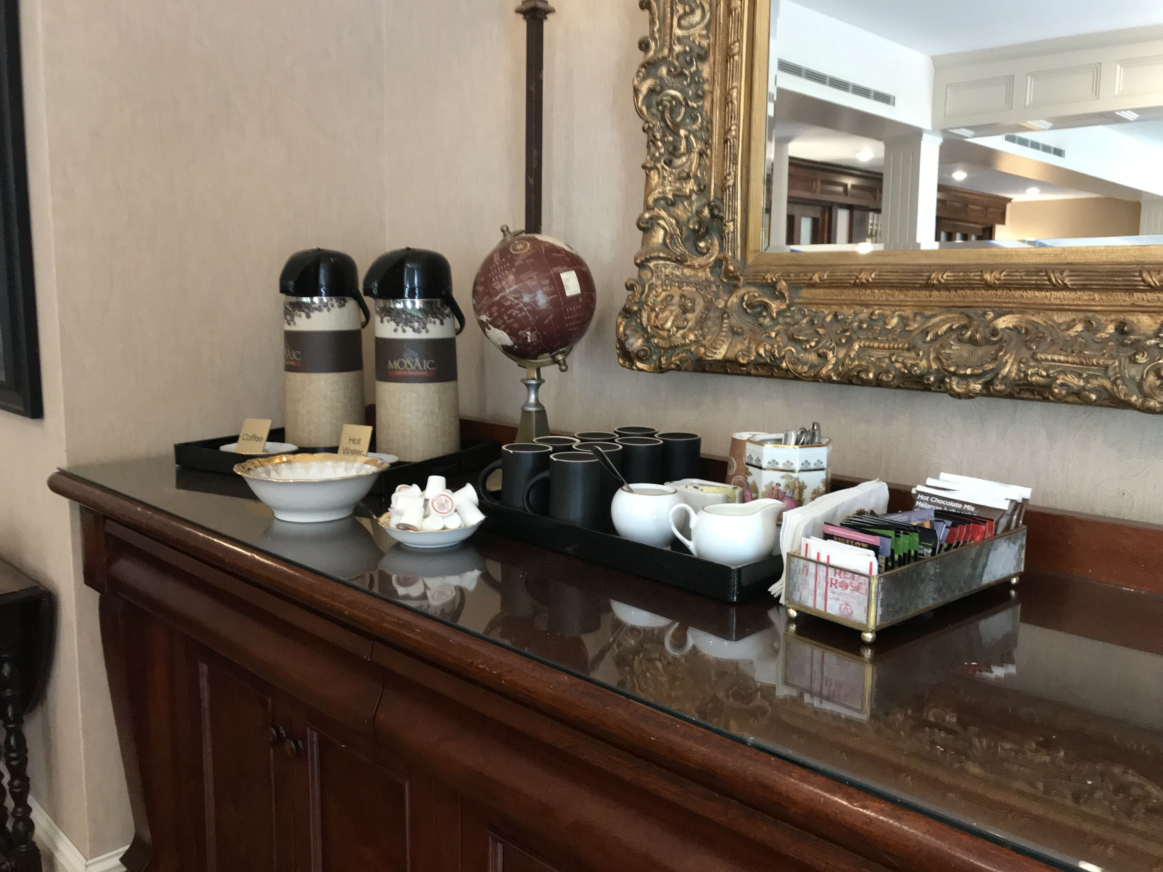 Un espace d’accueil d’hôtel avec un comptoir en verre où sont exposés des distributeurs de café et d  ’eau chaude, un globe terrestre et un plateau contenant des tasses et une petite boîte de chocolats.