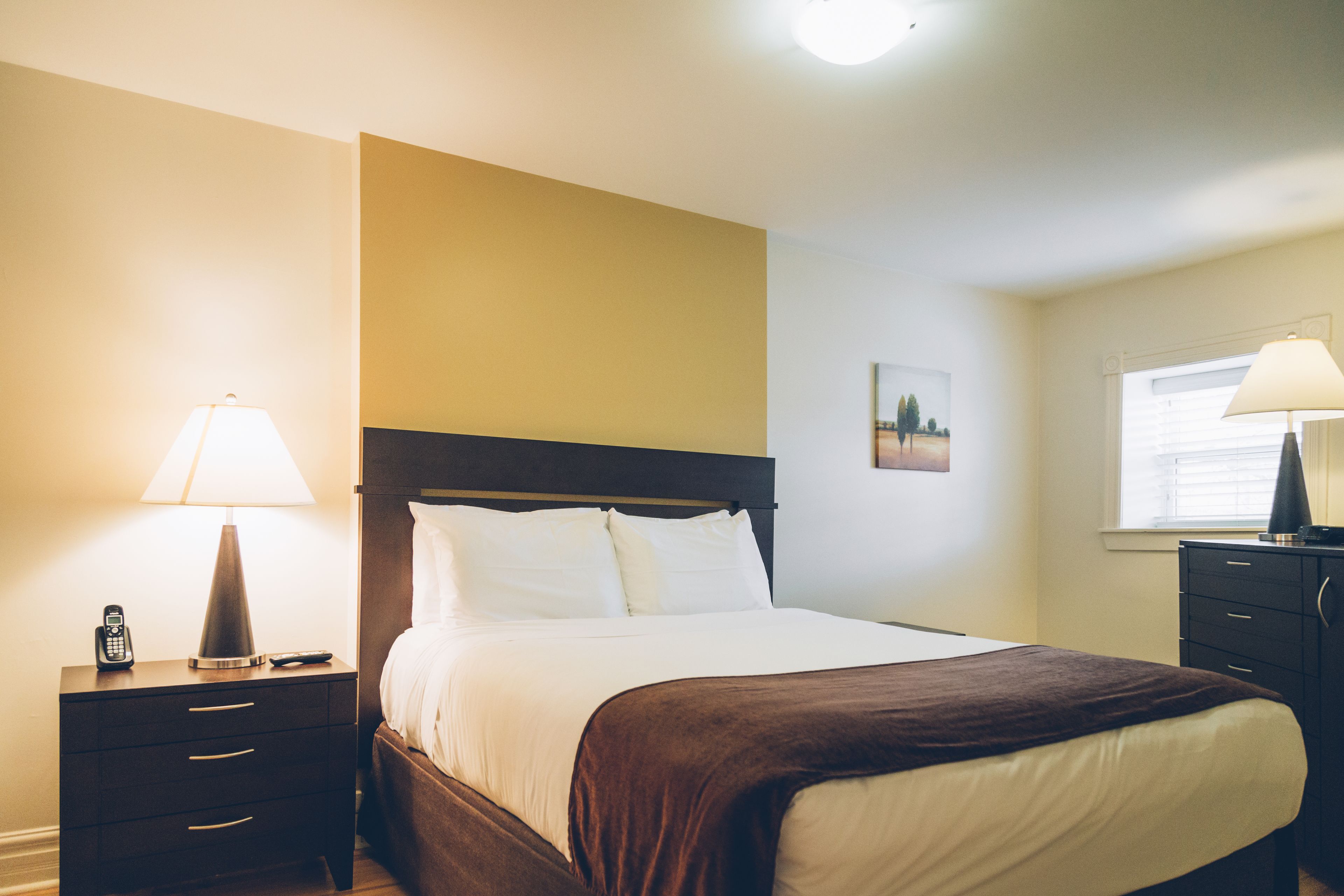 A hotel room with a bed, a nightstand with a lamp, a dresser, and a window with blinds.