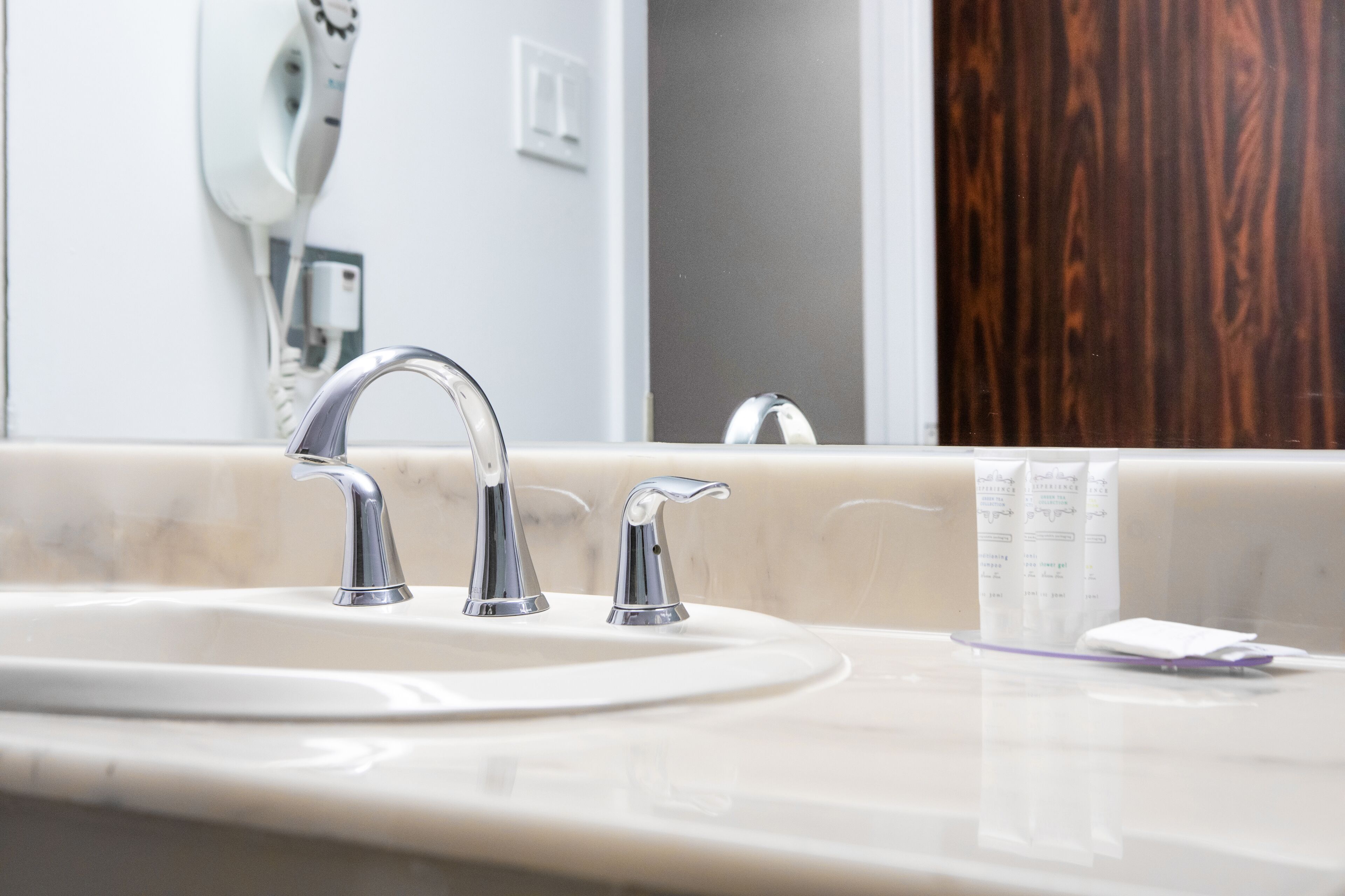 A bathroom sink with two faucets, a mirror, and a wooden door in the background.