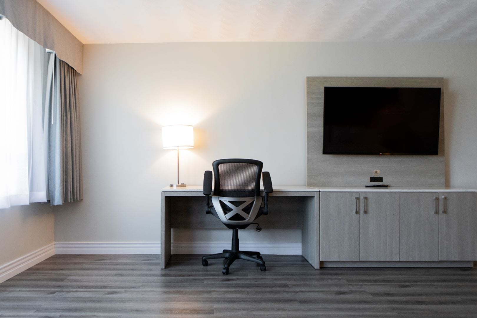 A hotel room with a desk, chair, TV, and a lamp.