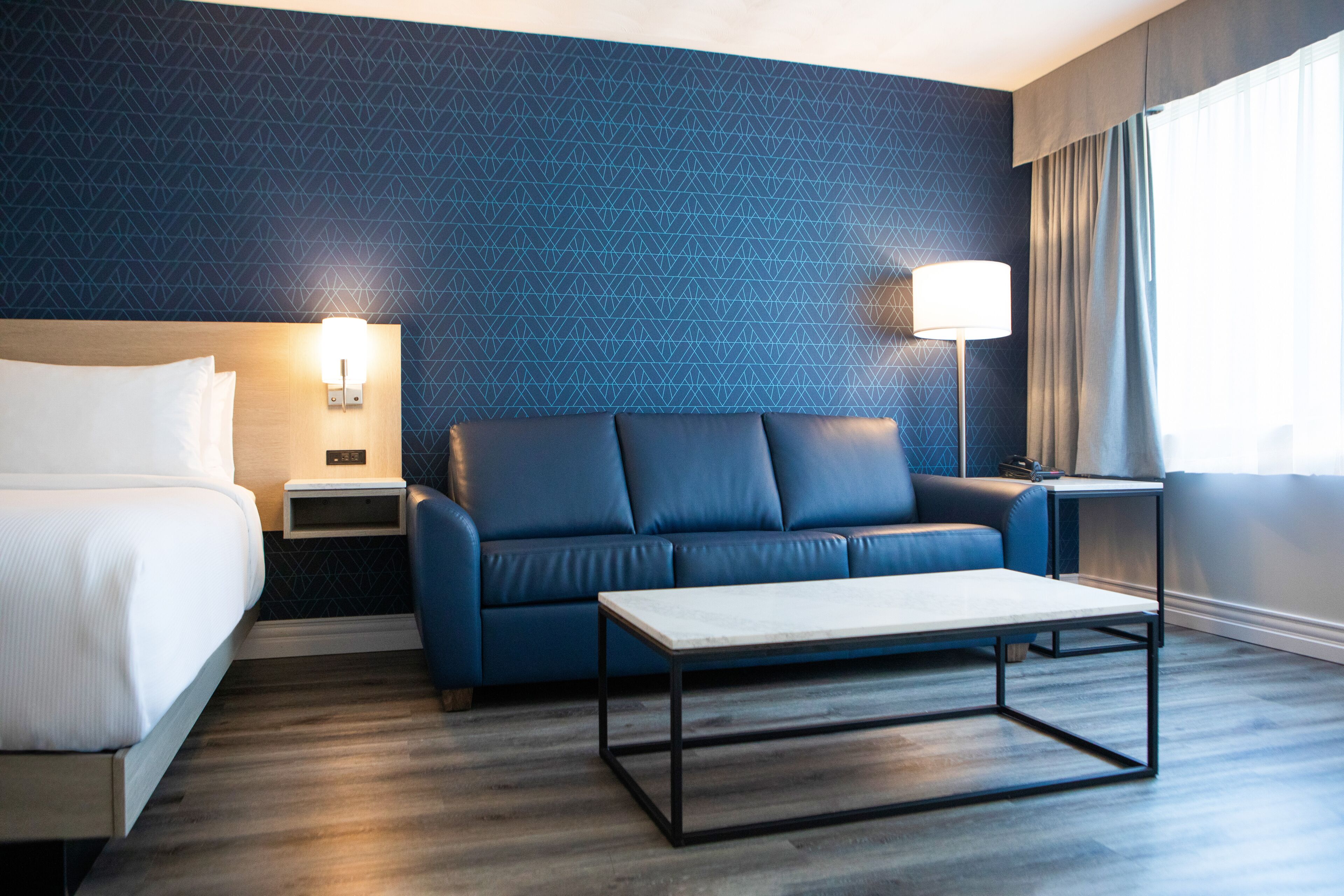 A hotel room with a bed, a blue sofa, a small table, a nightstand, and a lamp.