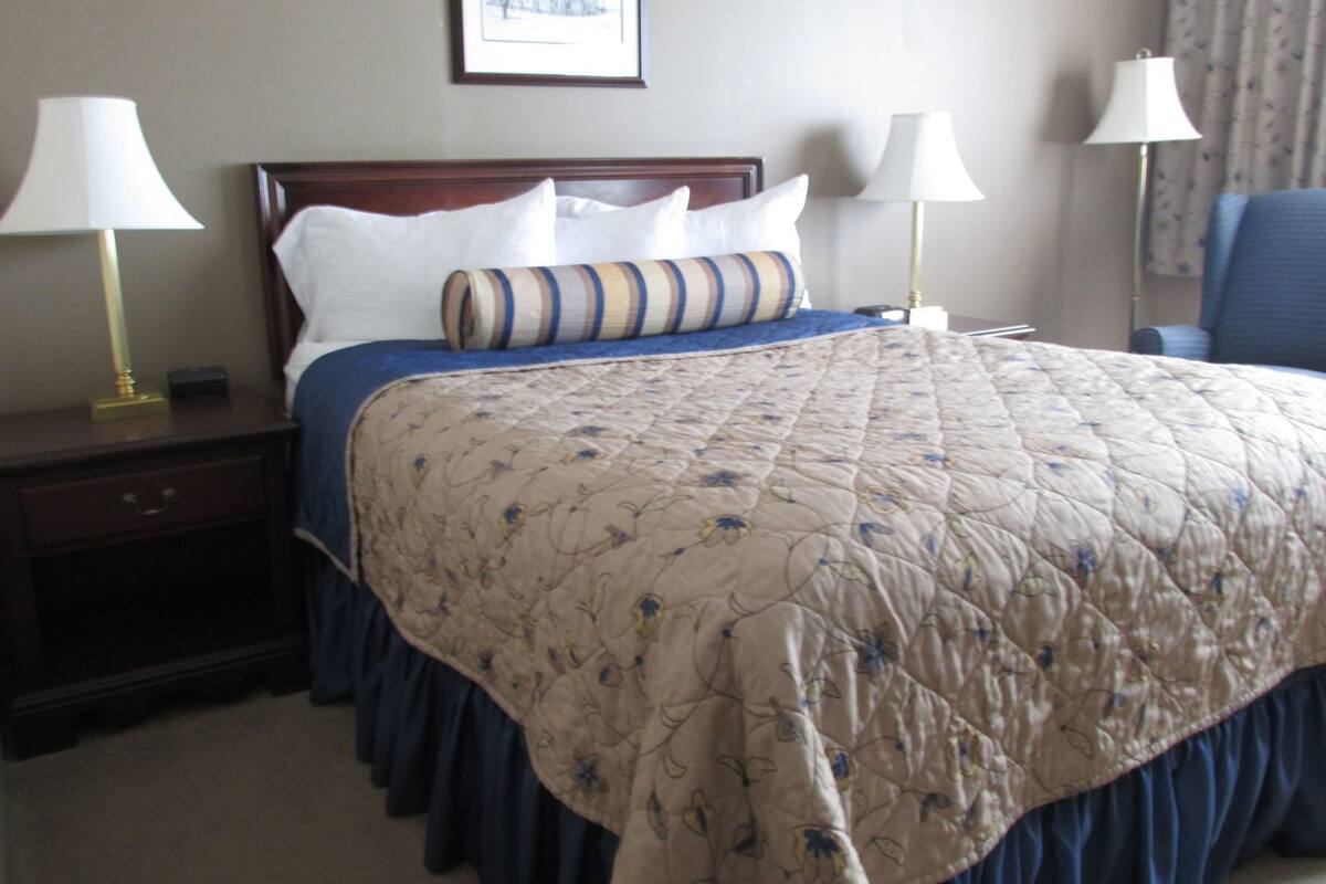 A neatly made bed with a floral quilt, two bedside lamps, a framed picture on the wall, and a chair in the corner.