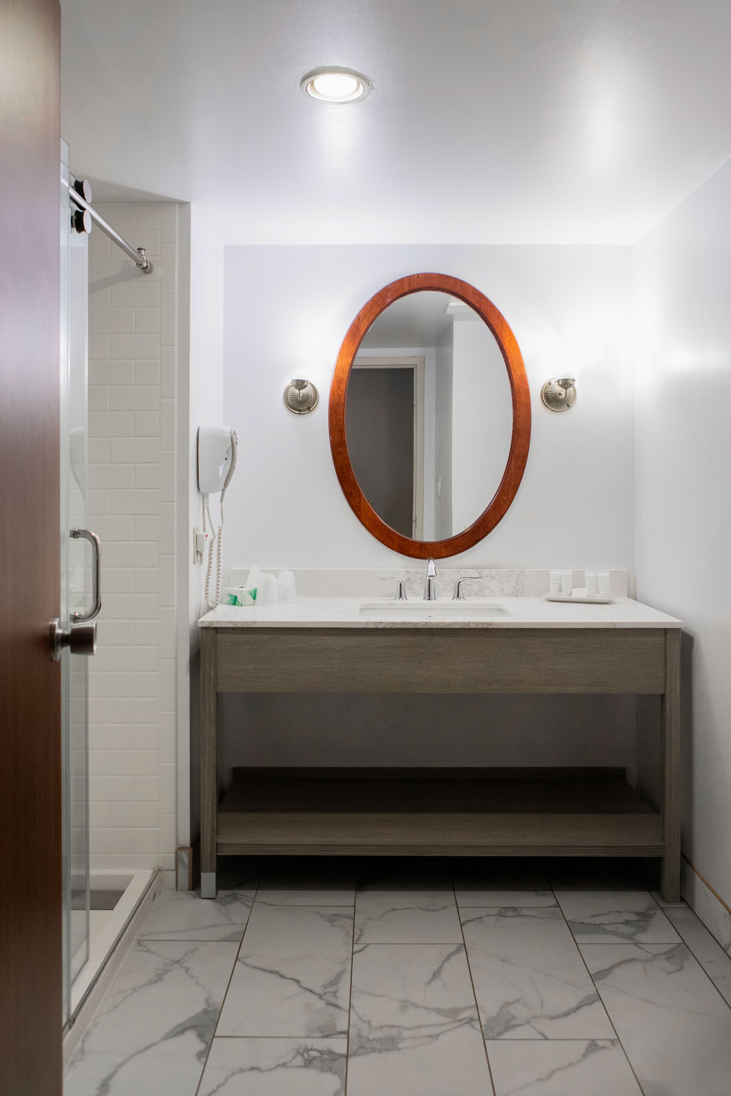 A bathroom with a wooden vanity, a round mirror, a handheld phone, and a shower area.