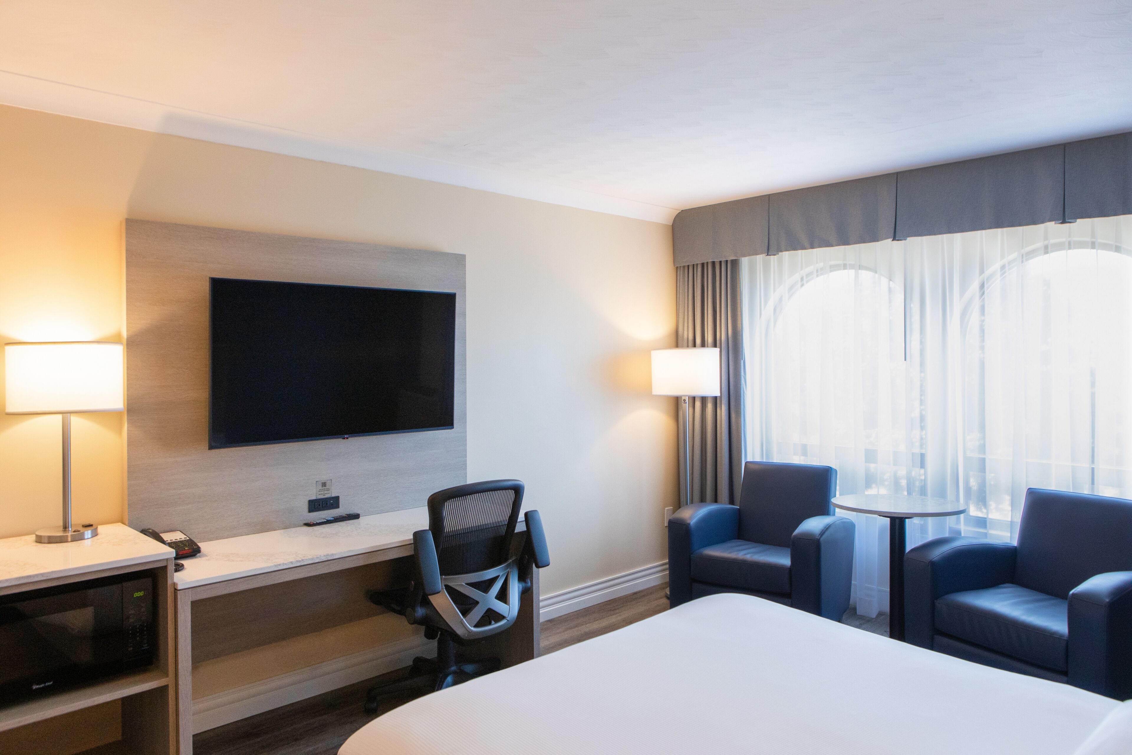 Habitación de hotel con cama, escritorio, silla, televisor, lámparas y ventana con cortinas.