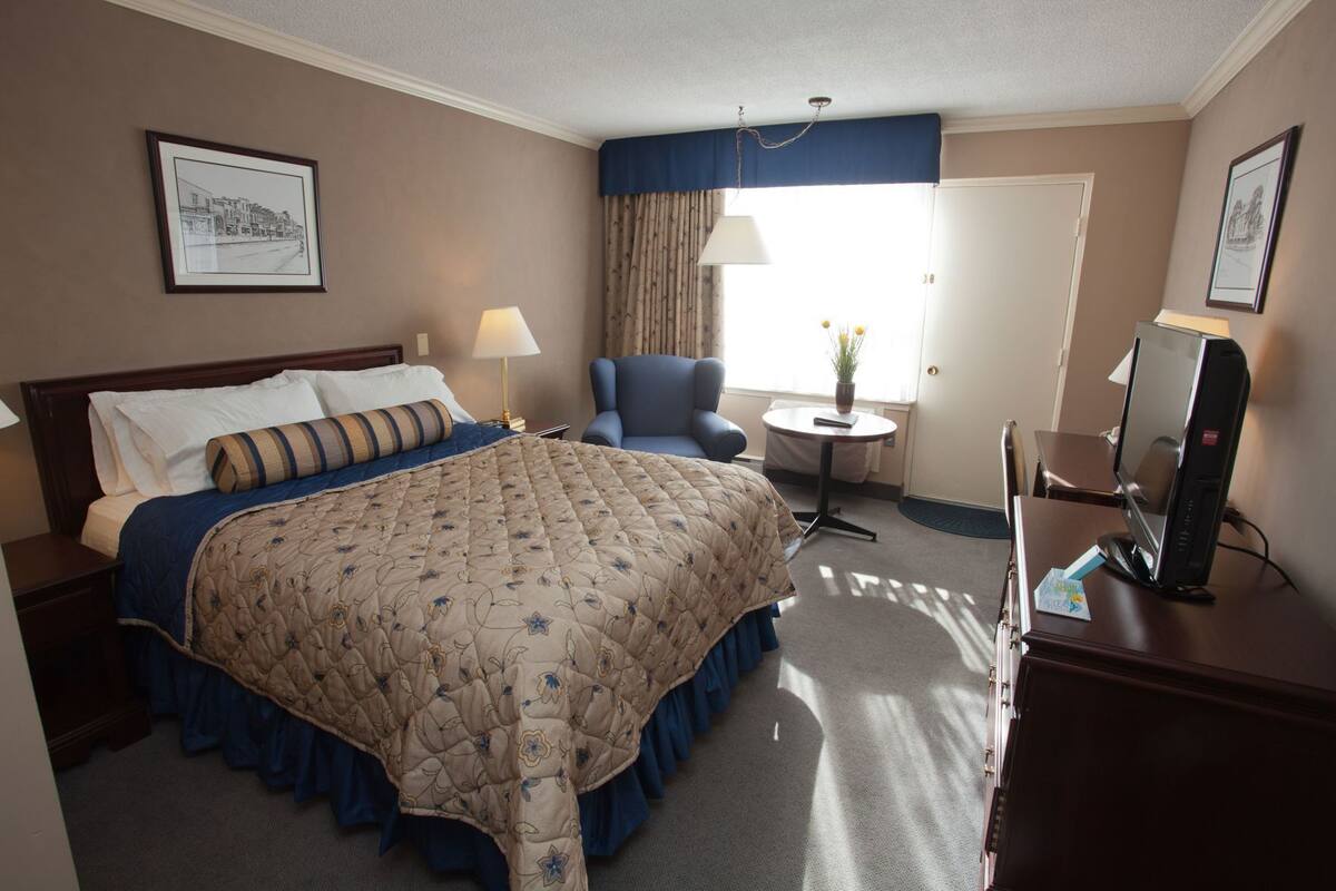 A hotel room with a large bed, a desk with a computer, a chair, a small table with a lamp, and a window with curtains.