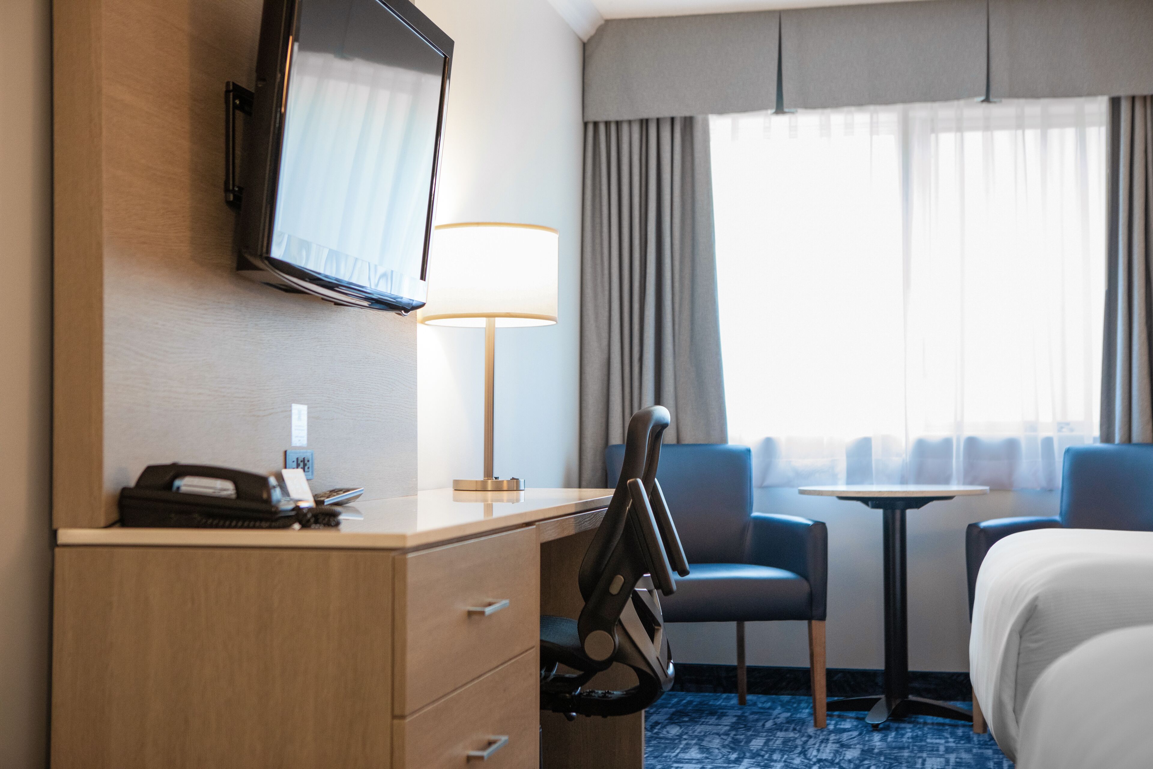 Quarto de hotel com cama, escrivaninha, cadeira, televisão, luminária e vista para o exterior pela janela.