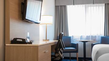 Quarto de hotel com cama, escrivaninha, cadeira, televisão, luminária e vista para o exterior pela janela.