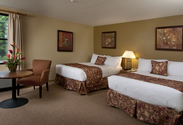 A hotel room with two beds, a chair, a table, and a lamp.